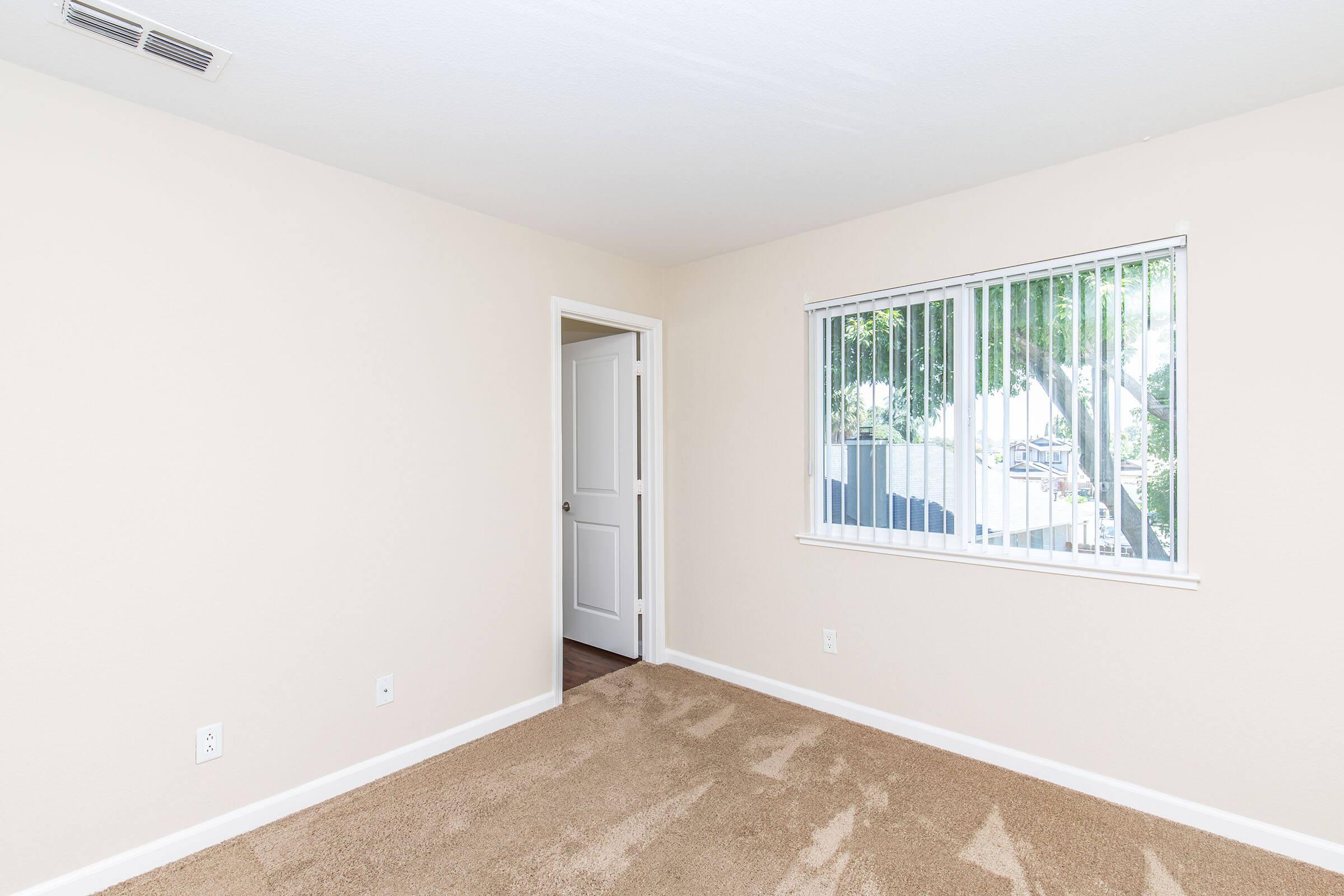 A vacant room with beige walls and light brown carpet. There is a closed door on the left leading to another space and a window with white blinds on the right, allowing natural light to enter. The room is empty, giving a sense of openness and potential for furniture placement.