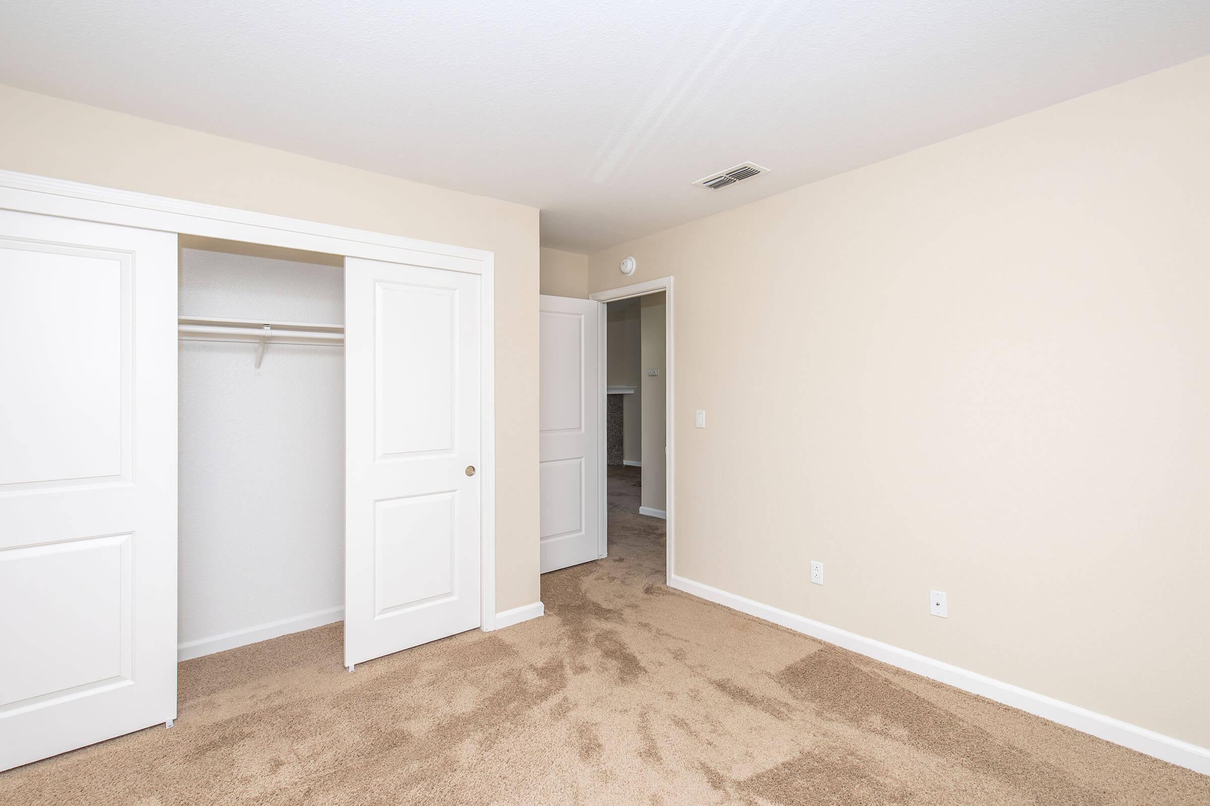 Empty room with beige walls and carpeted flooring. There is a closet with white double doors on the left side. A second door leads to another space, partially visible in the background. The overall appearance is clean and bright, suggesting a modern living area.