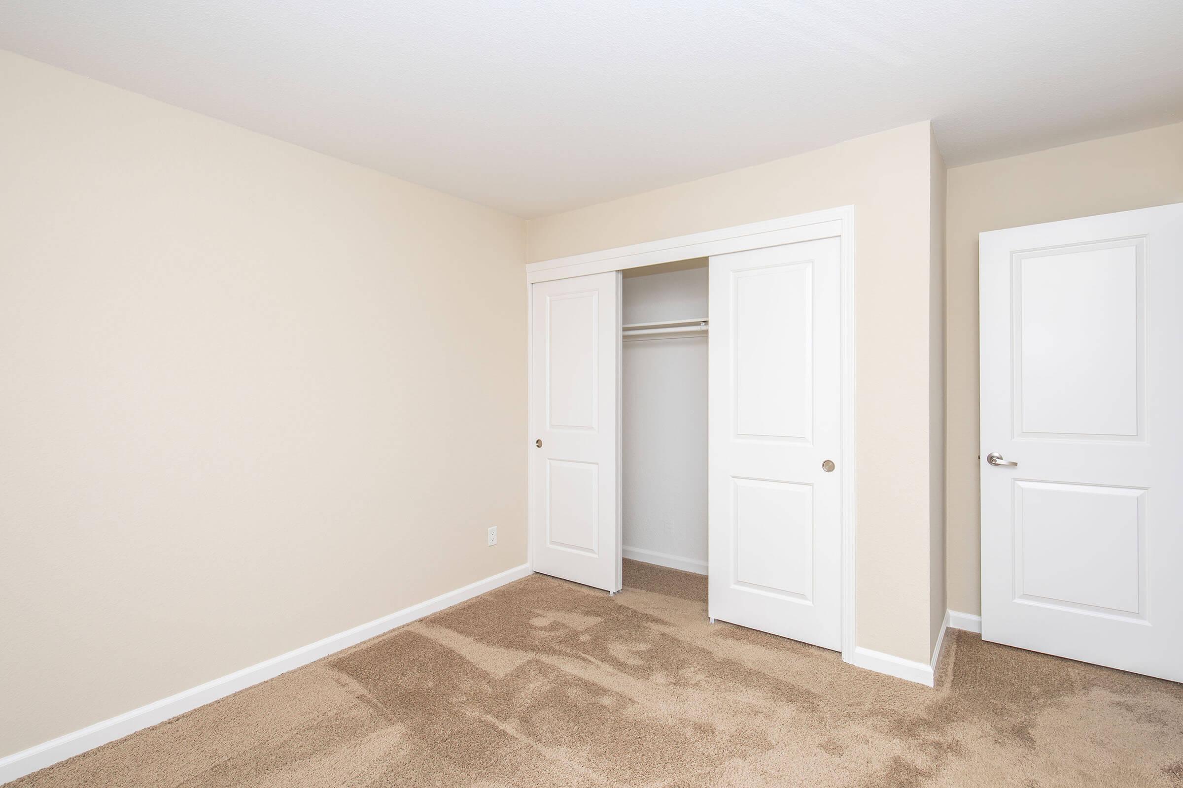 Empty room with neutral beige walls and carpet. A closet with double doors is visible on the left, and a closed door is present on the right. The room is well-lit and unfurnished, creating a spacious feel.