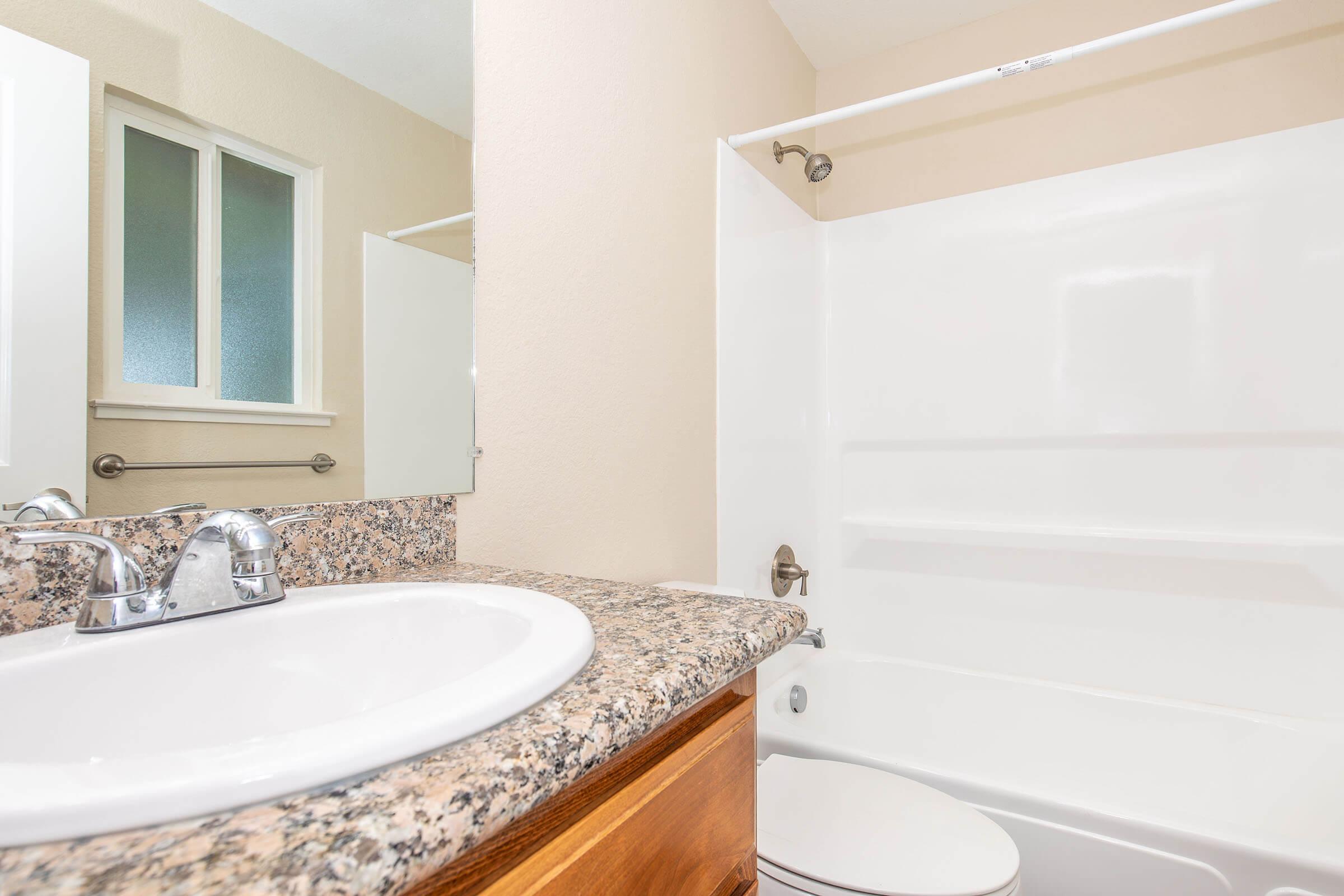 A clean, modern bathroom featuring a sink with a granite countertop, a white bathtub and shower unit, a wooden cabinet, and a mirror above the sink. Natural light comes through a window, creating a bright and inviting atmosphere.
