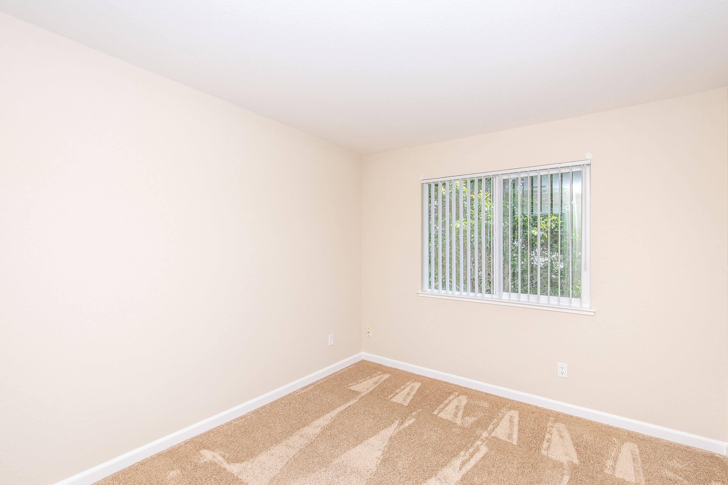 A simple, unfurnished room with light beige walls and a window featuring vertical blinds. The floor is covered in light-colored carpet. Soft shadows are visible on the carpet, likely from objects outside the window. The overall atmosphere is bright and minimalist.