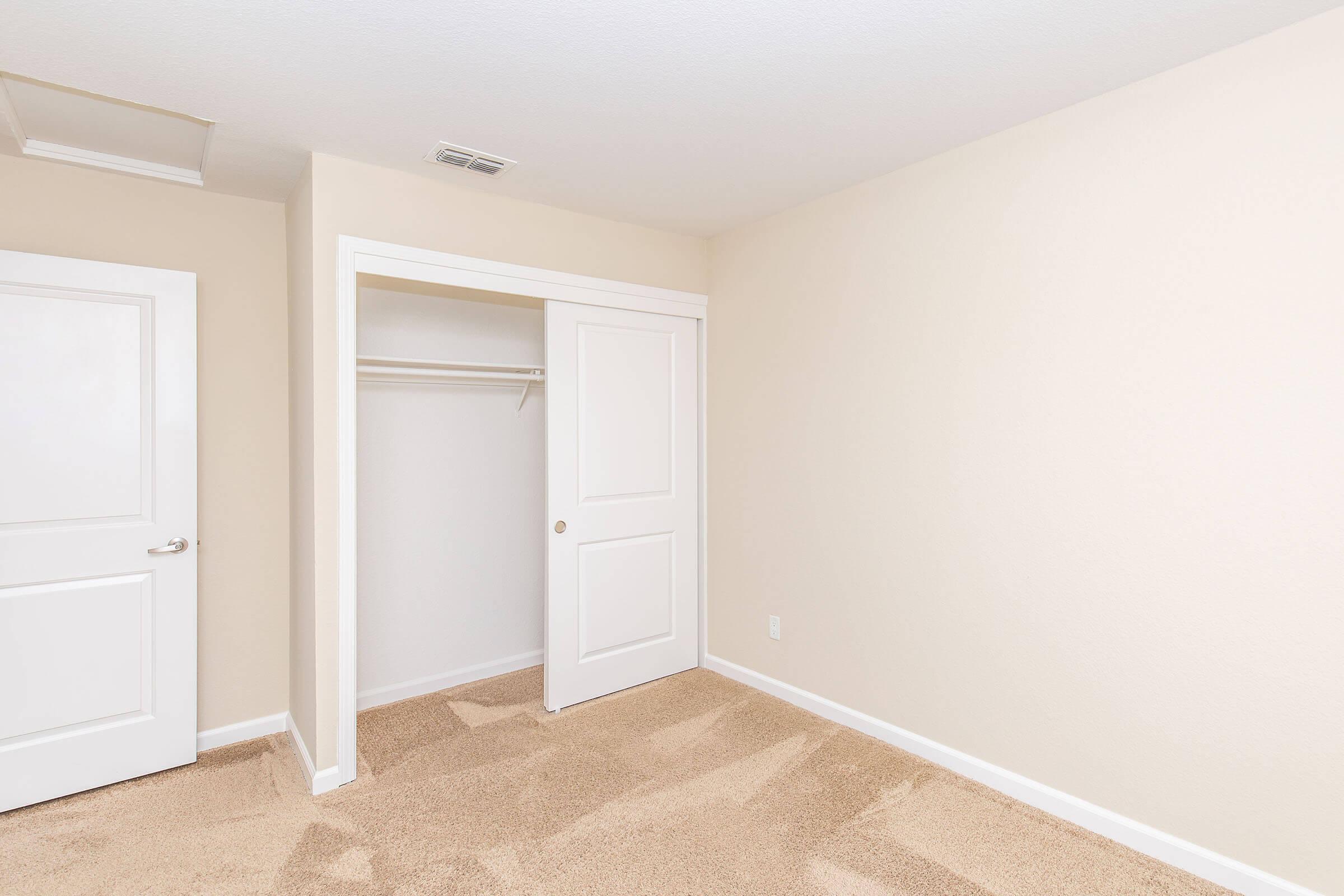 Empty room with light beige walls and carpeted floor. There are two white doors, one leading to a closet with a hanging rod and no shelves. The room is well-lit, and the overall space appears clean and ready for furnishing.