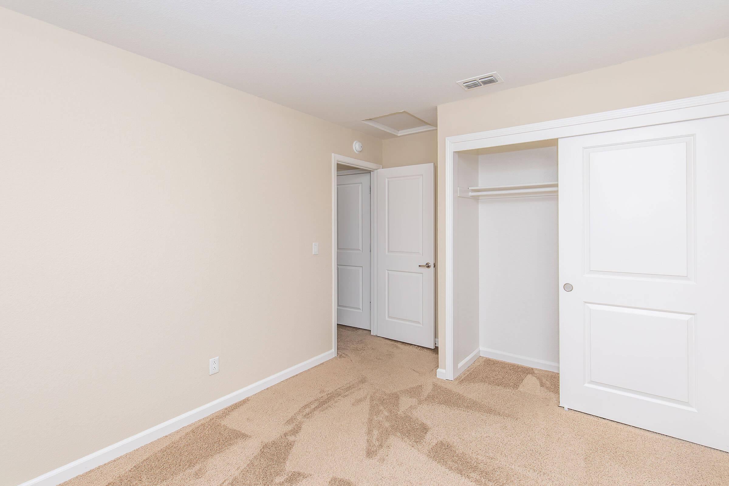 A small, empty room with light beige walls and carpet. There are two doors on the left leading to adjacent spaces and a closet with a sliding door. The room has minimal furnishings, giving it a clean and spacious appearance. Bright and well-lit with natural light.
