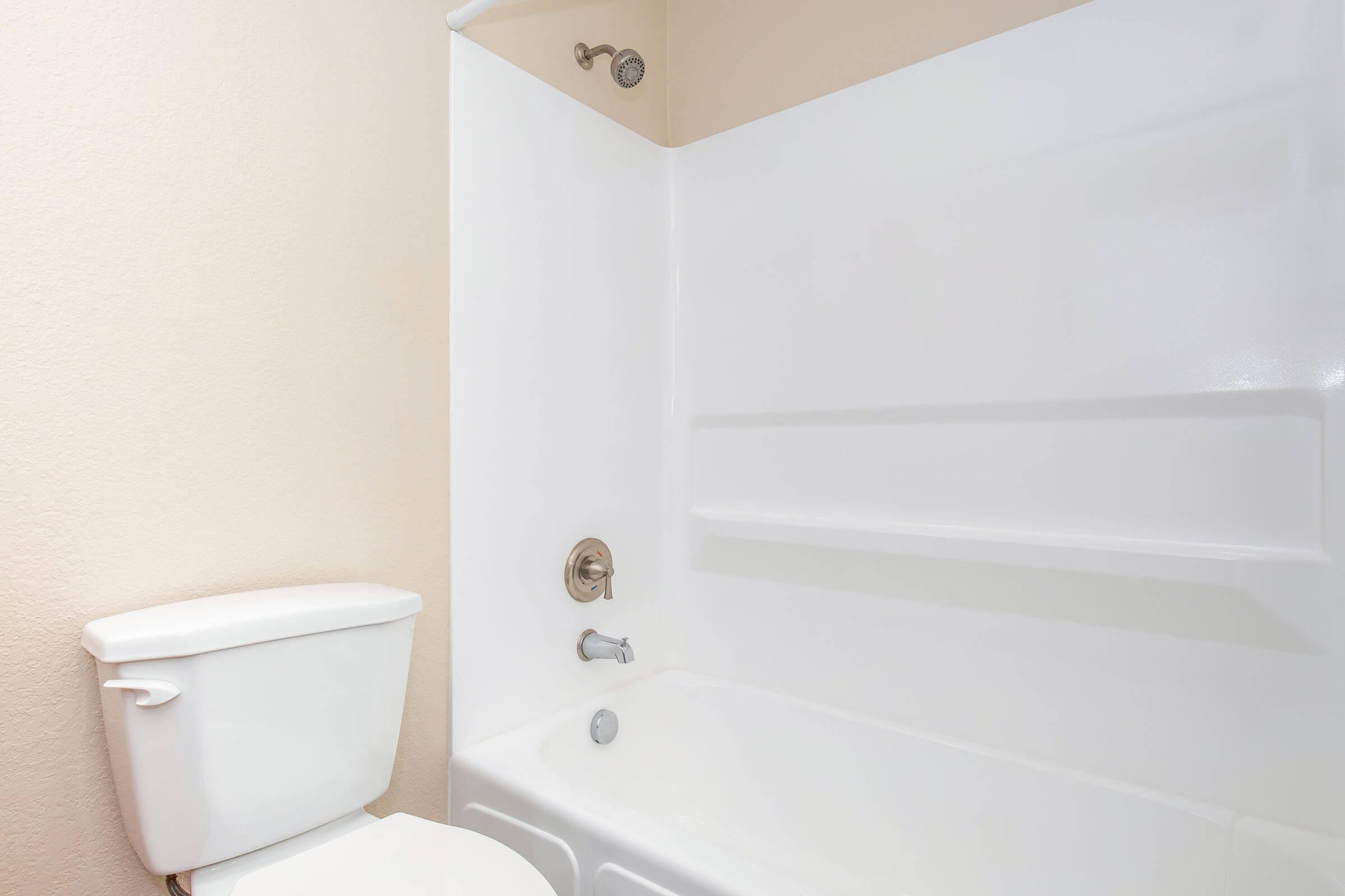 A clean, modern bathroom featuring a white bathtub with a showerhead, a wall-mounted faucet, and a white toilet. The walls are a light color, and there are built-in shelves in the bathtub area. The overall design is minimalist and well-maintained.
