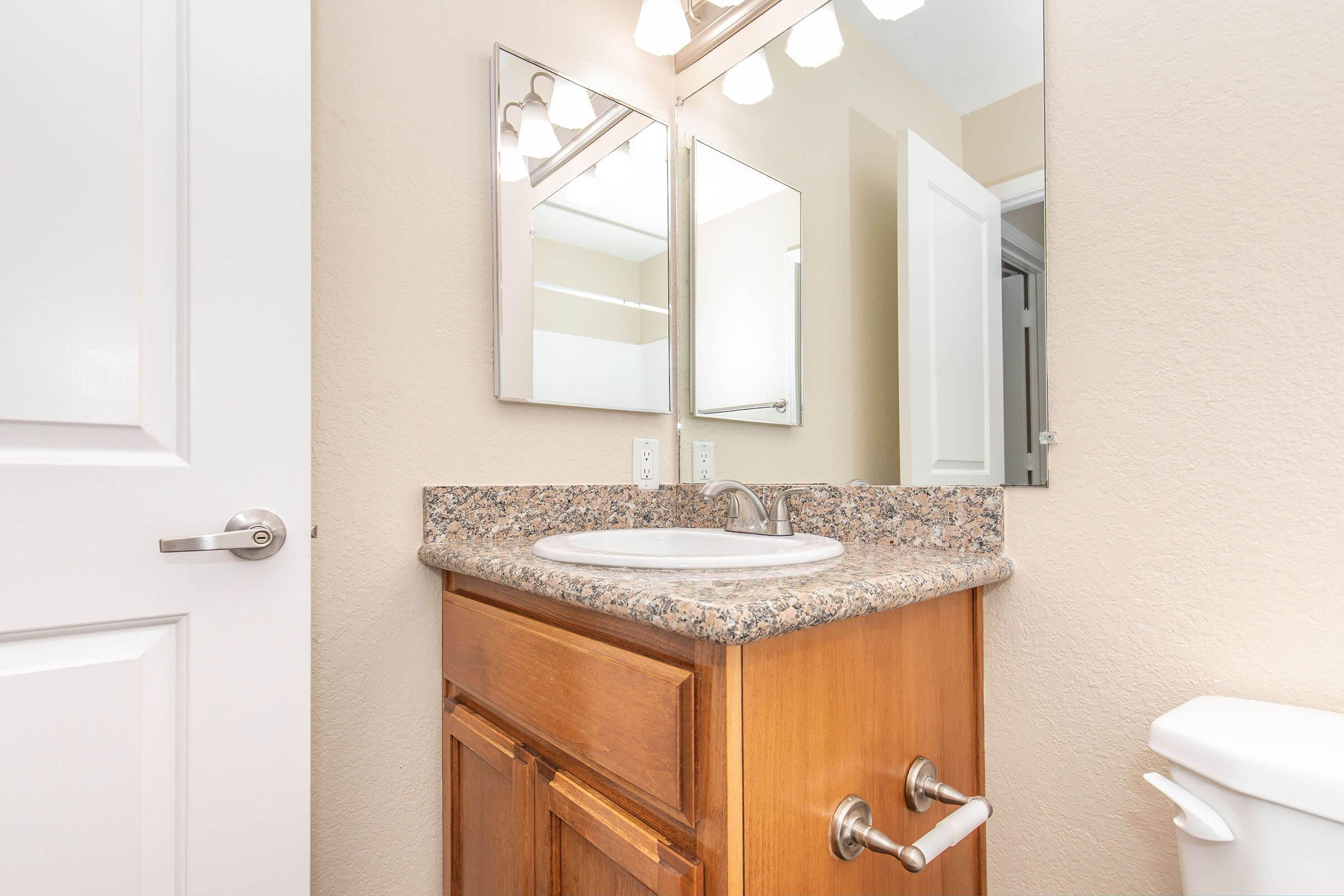 A clean bathroom featuring a wooden vanity with a granite countertop, a white sink, and two rectangular mirrors hanging above it. A light fixture is mounted above the mirrors, and a white toilet is visible to the side. The walls are painted a light color, creating a bright atmosphere.
