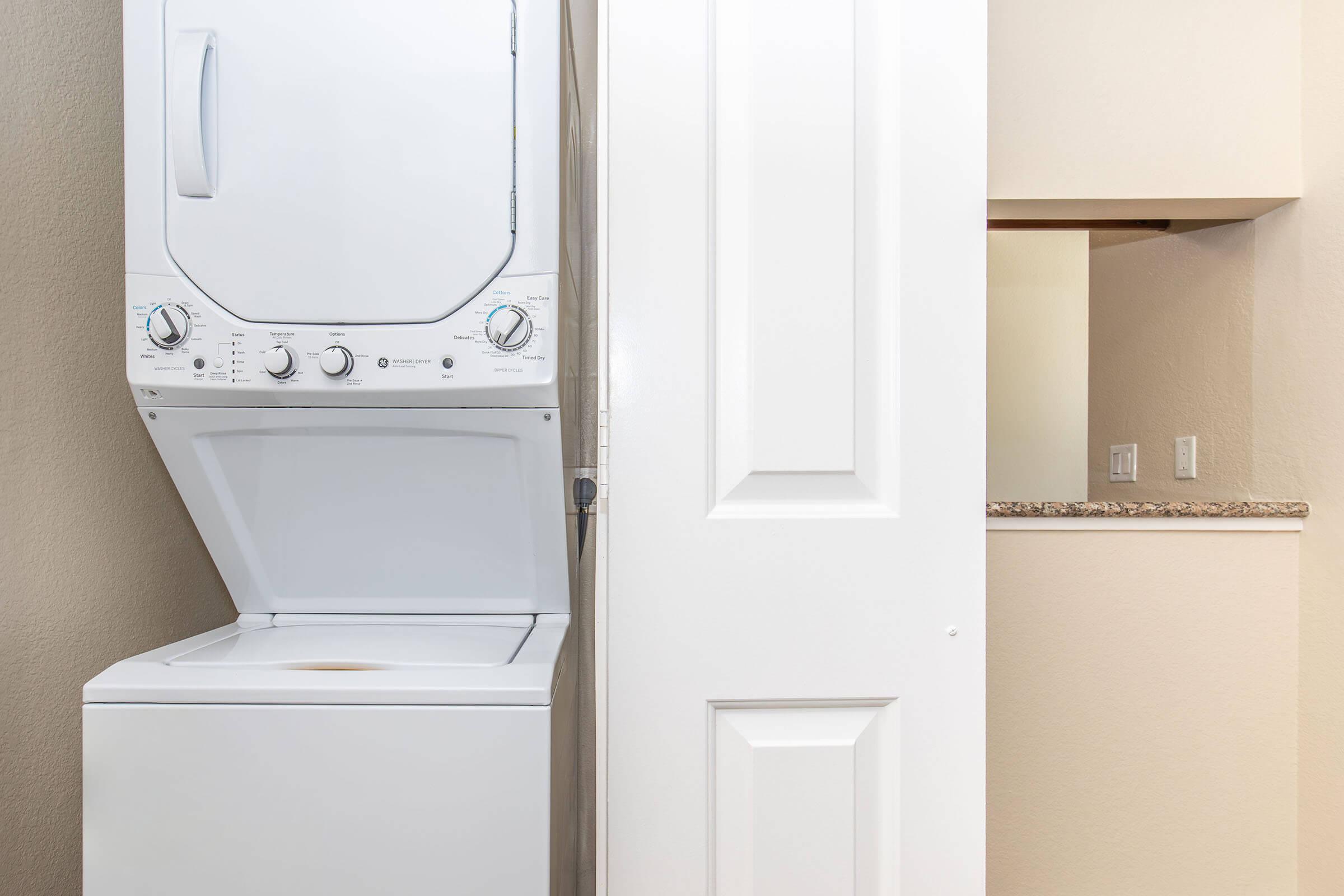 A stacked washer and dryer unit is positioned next to a closed white door, with a small opening to a kitchenette visible in the background. The walls are painted a light color, creating a clean and organized space.