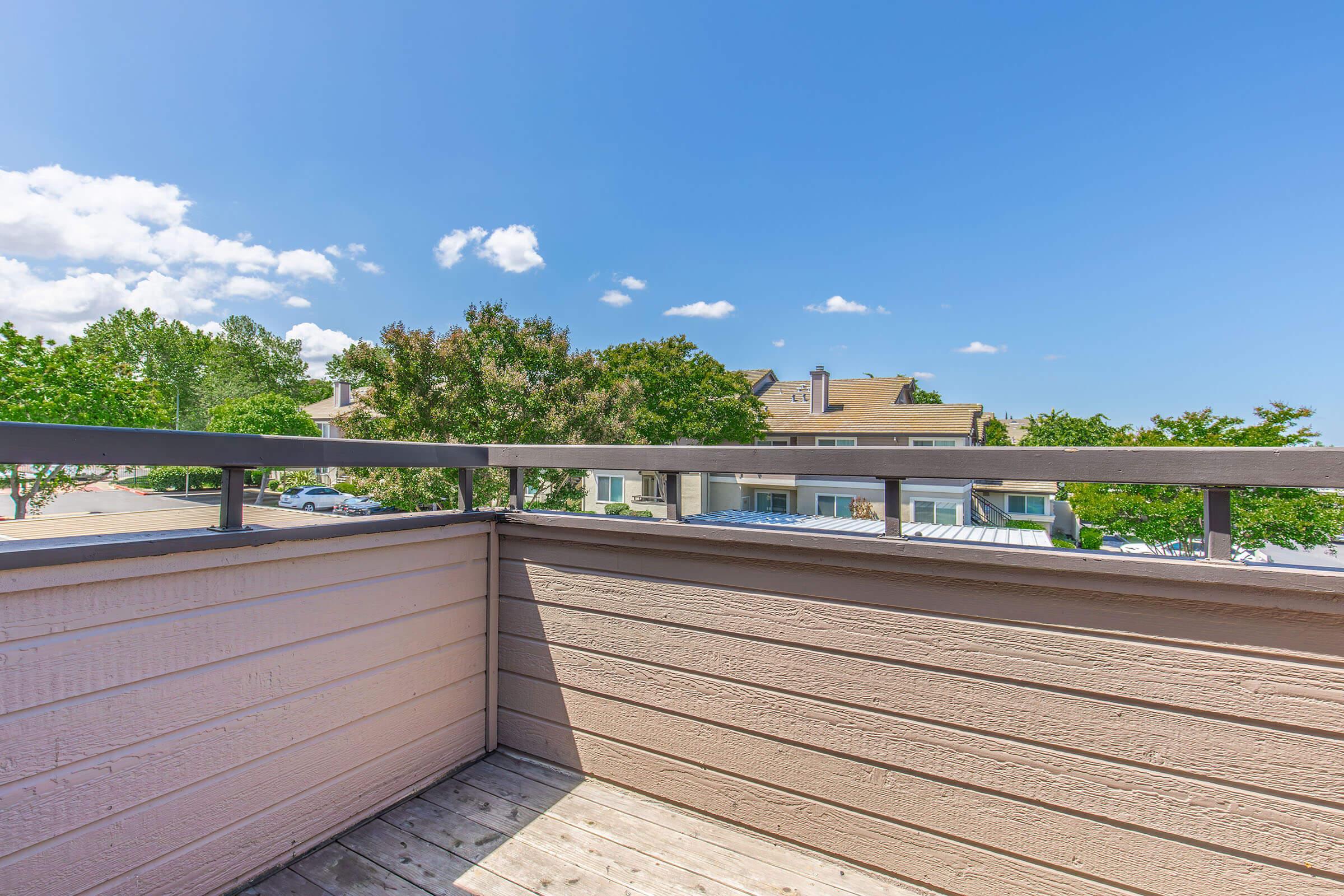 A balcony view featuring a wooden railing and a wooden deck, overlooking a neighborhood with trees and residential buildings under a clear blue sky with a few clouds.