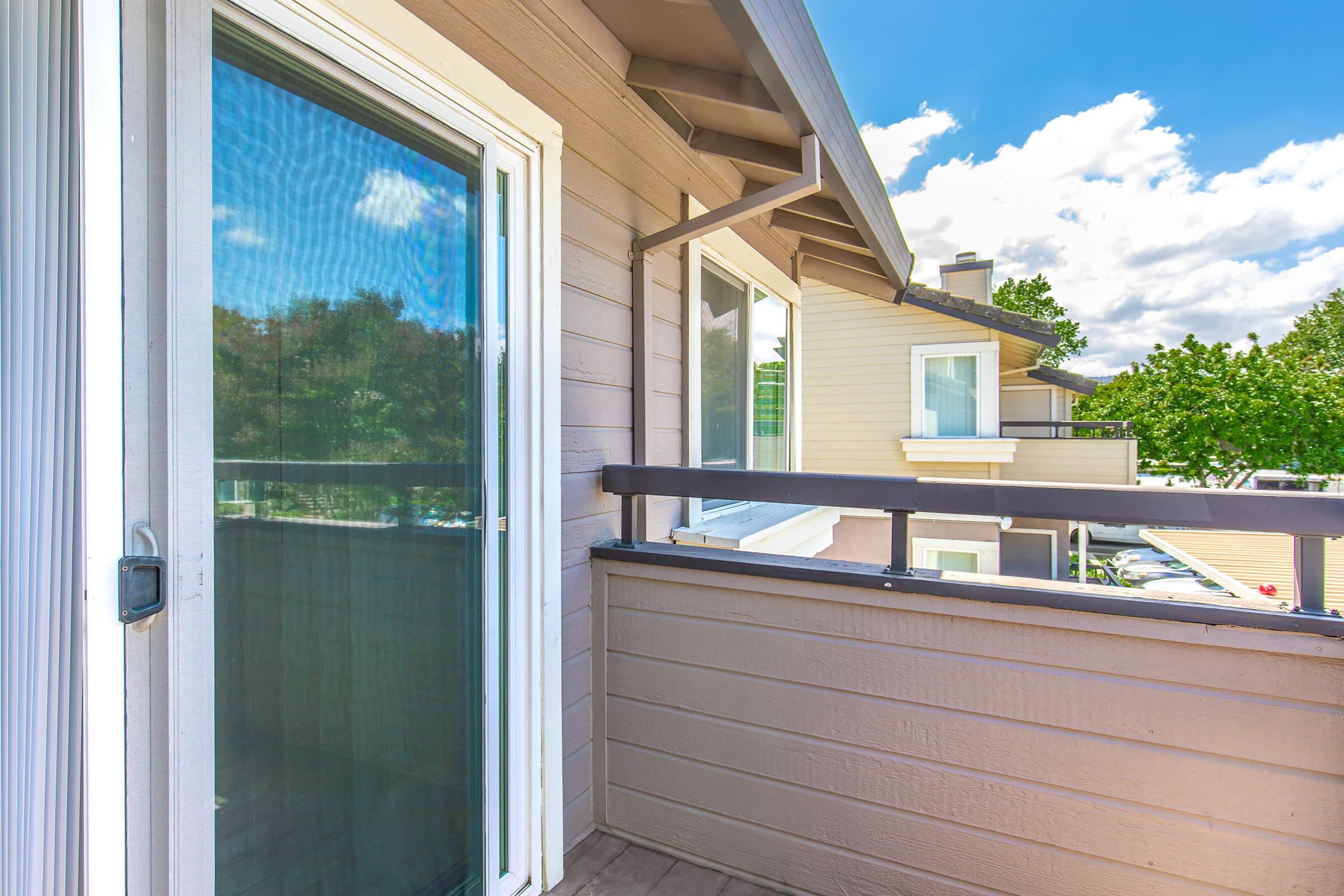 View of a balcony with a sliding glass door, featuring a wooden railing. The exterior walls are light-colored, and there's a clear blue sky with fluffy white clouds. Trees can be seen in the background, adding a touch of nature to the urban setting. The scene conveys a sense of openness and tranquility.