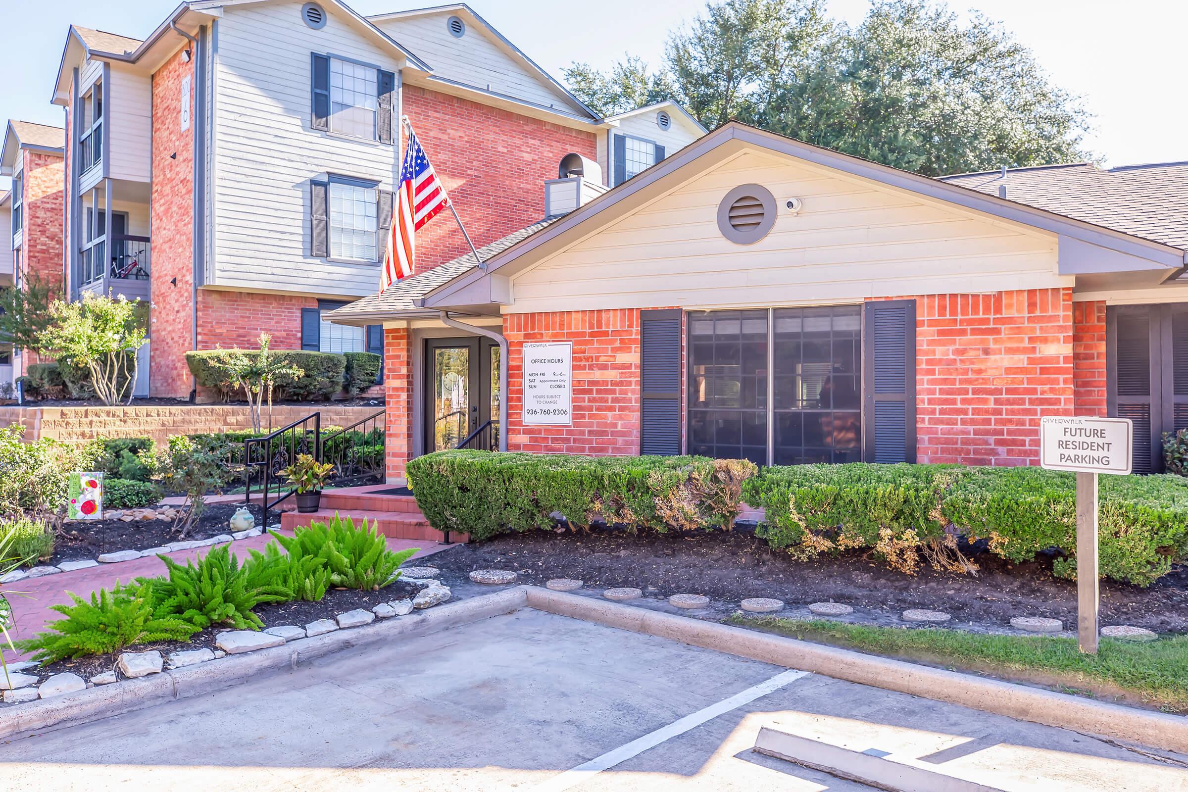 A brick building with a welcoming entrance, featuring an American flag. Lush green landscaping surrounds the area, and a sign indicates future resident parking. In the background, there are additional multi-story apartment buildings under clear blue skies.