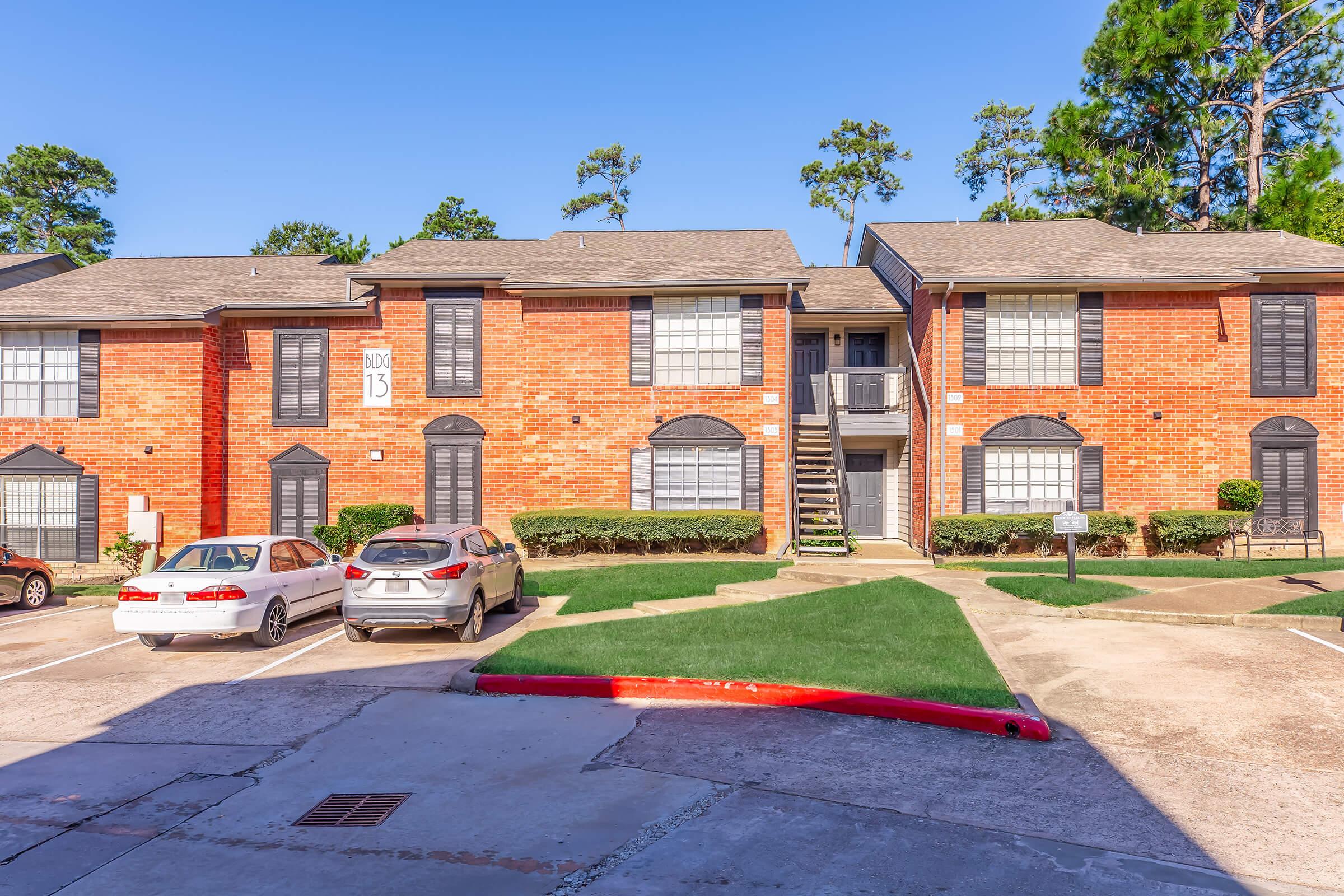 A two-story brick apartment building with multiple units, featuring landscaped grassy areas and a parking lot. The building has stairs leading to the second floor and a sign with the number 13. Clear blue sky and surrounding trees are visible.
