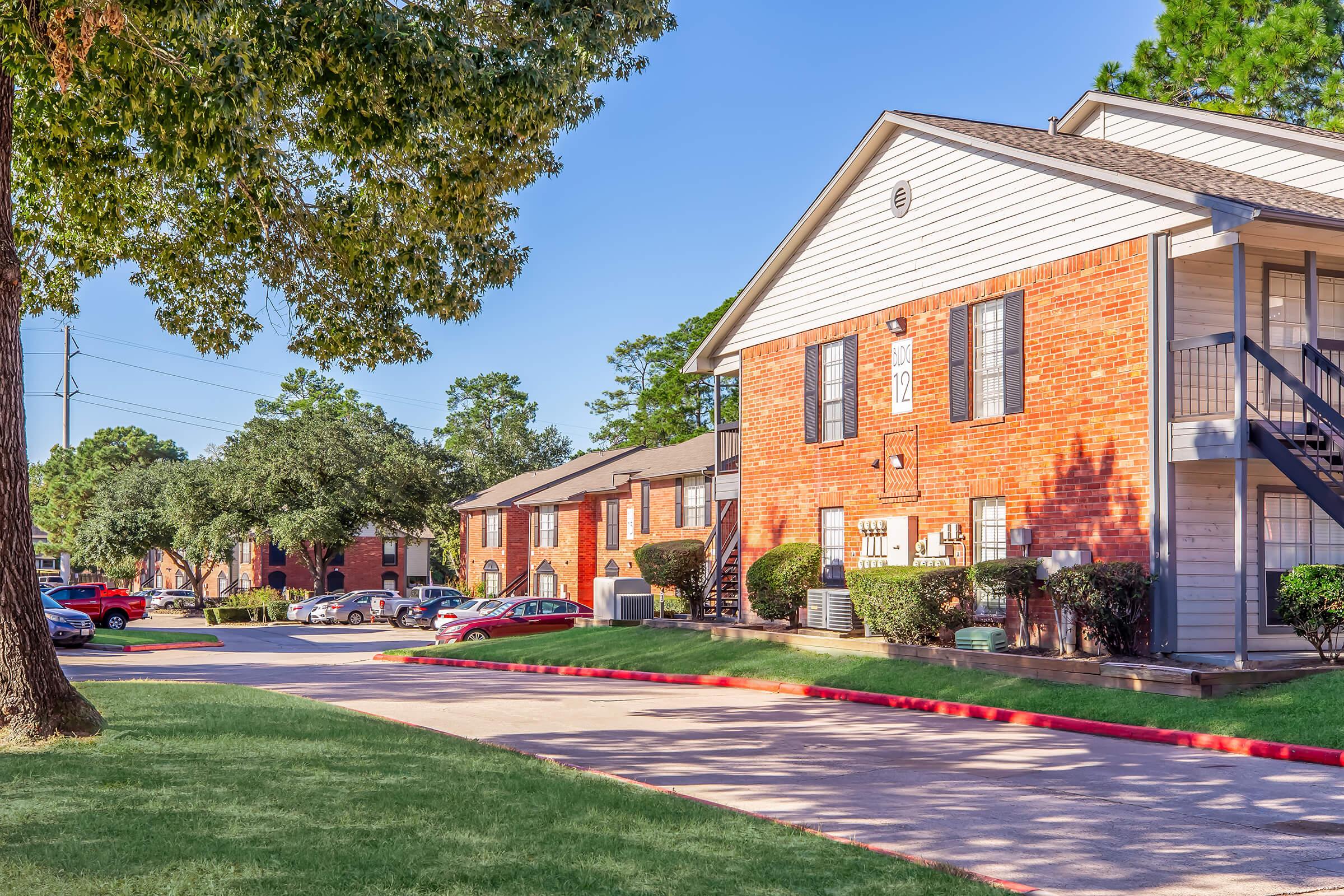 A well-maintained residential complex featuring brick buildings with white trim, surrounded by green grass and trees. The scene shows a paved driveway with parking spaces, along with several parked cars, creating a welcoming neighborhood atmosphere under a clear blue sky.