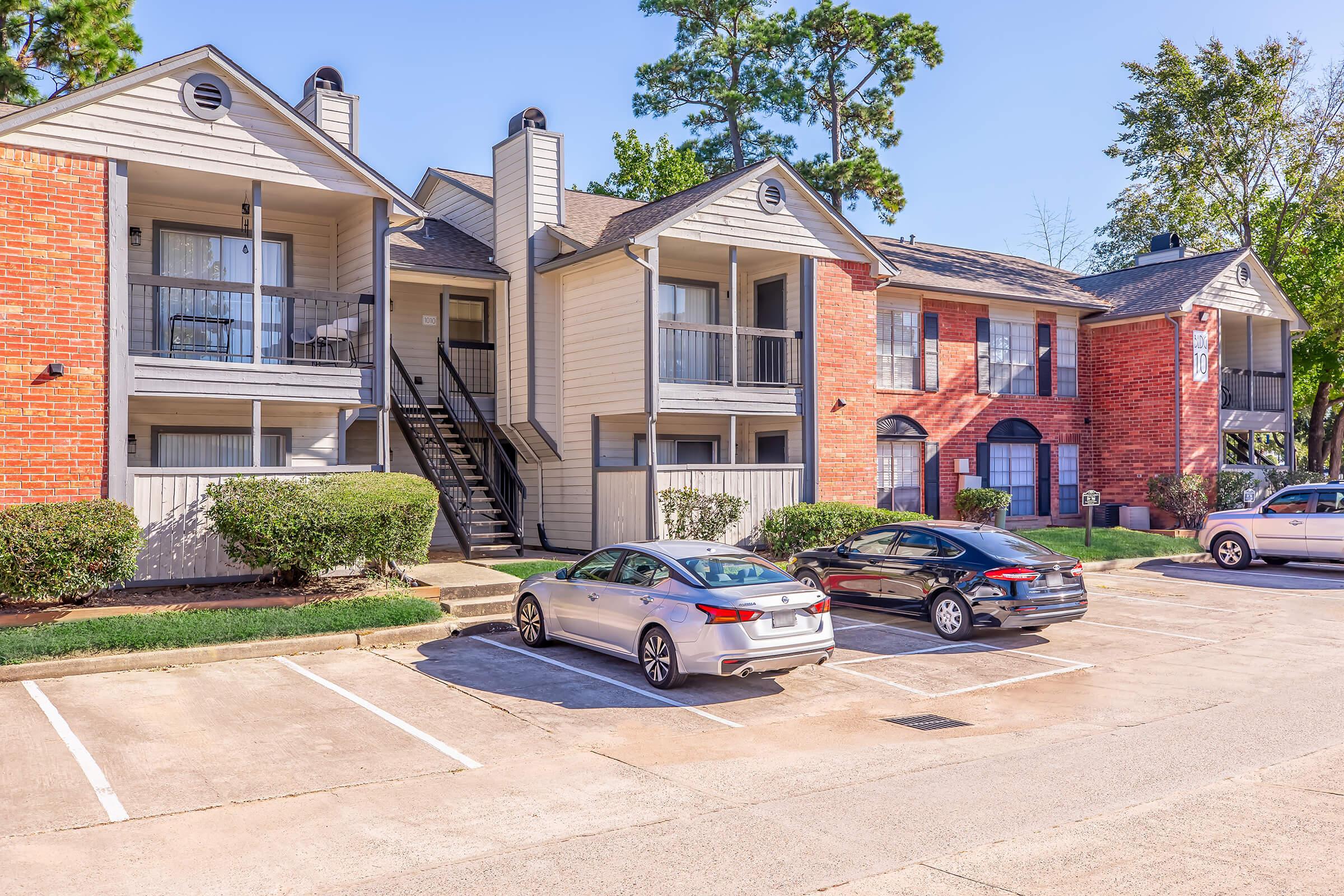 A view of a residential apartment complex featuring two buildings with a mix of brick and siding exteriors. There are several parked cars in the foreground and well-maintained landscaping with shrubs. The scene is set on a clear day with blue skies and trees in the background.