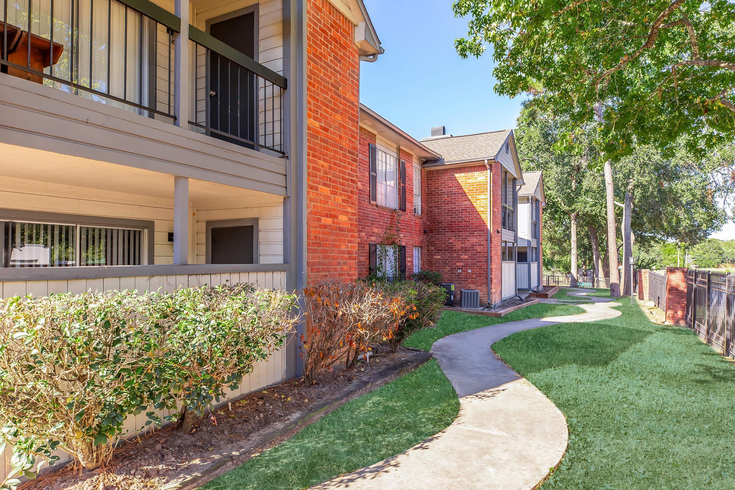 Apartment complex exterior featuring a path winding through neatly landscaped greenery, with a mix of brick and wooden balconies. The sky is clear and blue, and sunlight casts shadows on the walkway. The area appears well-maintained and inviting.