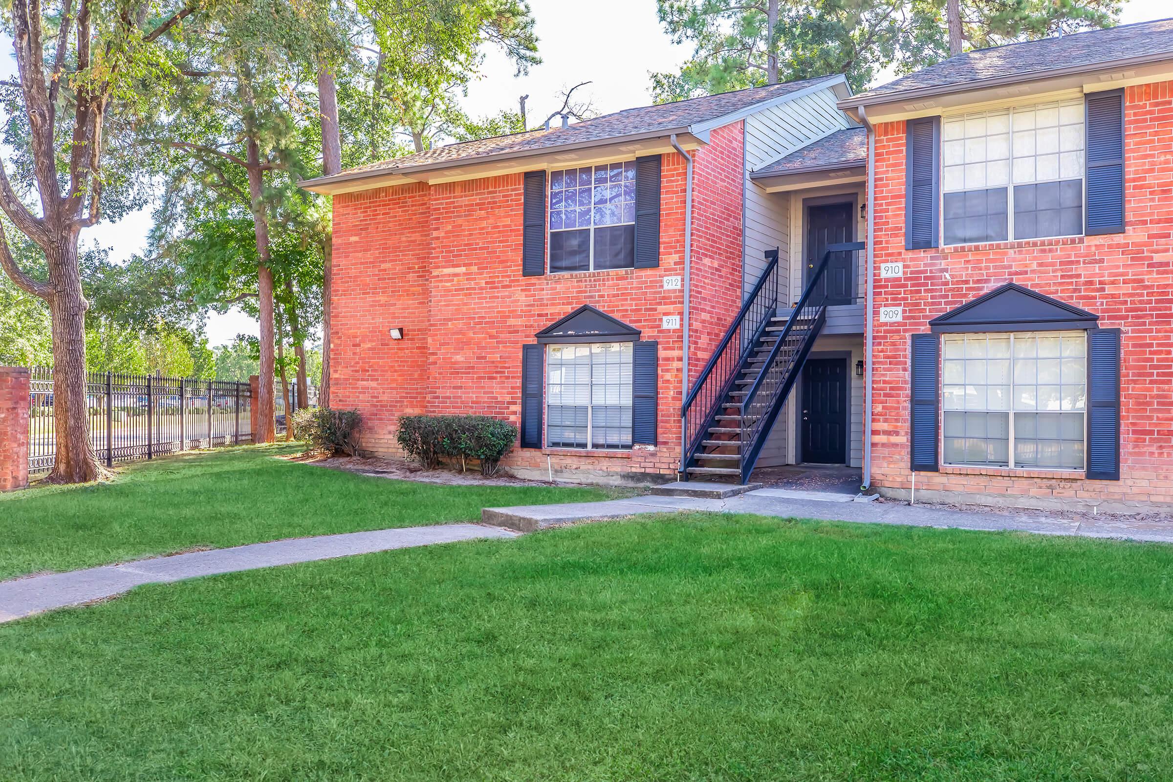 A brick apartment building featuring two floors, with light-colored trim and black shutters. A landscaped area with green grass and small bushes is in the foreground. A concrete pathway leads up to the entrance, with a set of stairs visible on the right side. Trees are present in the surroundings.