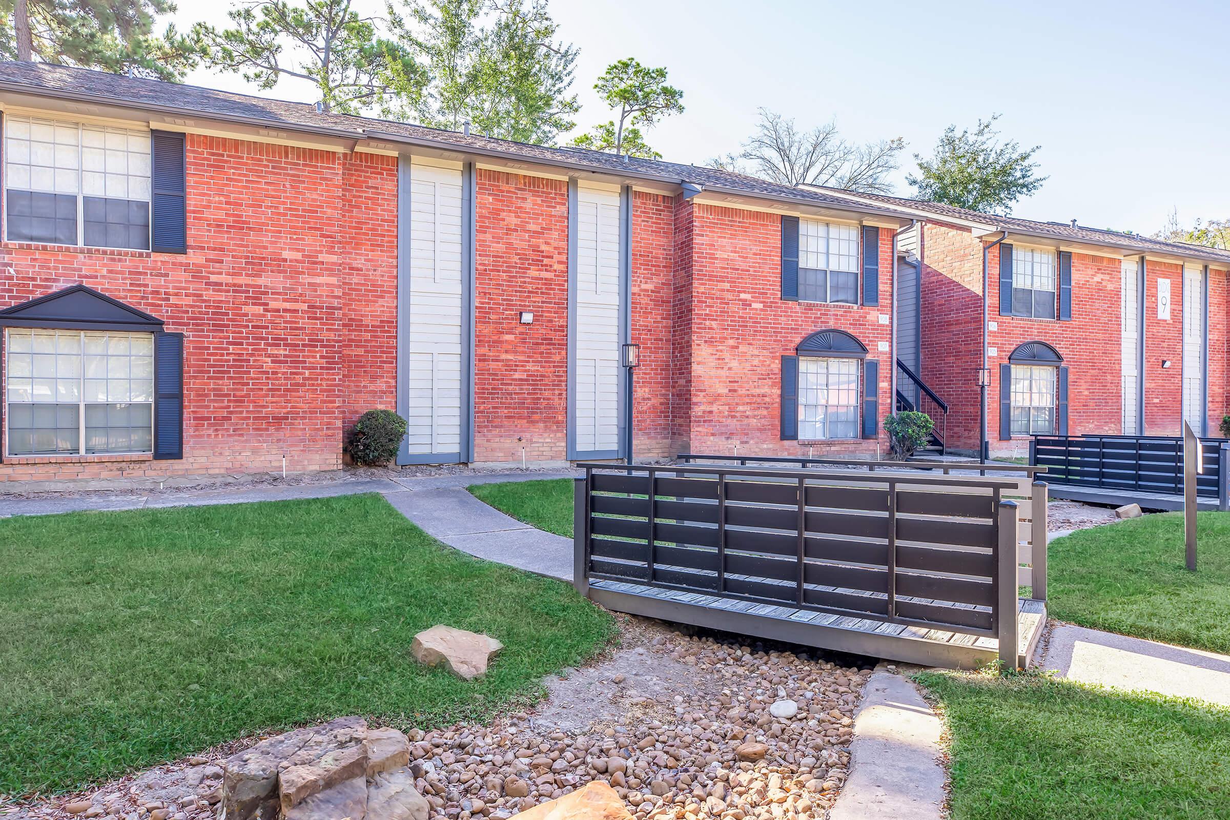 A view of a brick apartment building with multiple units, featuring large windows and black railings. A paved walkway leads to a small wooden bridge over a gravel path, surrounded by manicured grass and shrubs. Sunlight illuminates the scene, highlighting the architecture and landscaping.