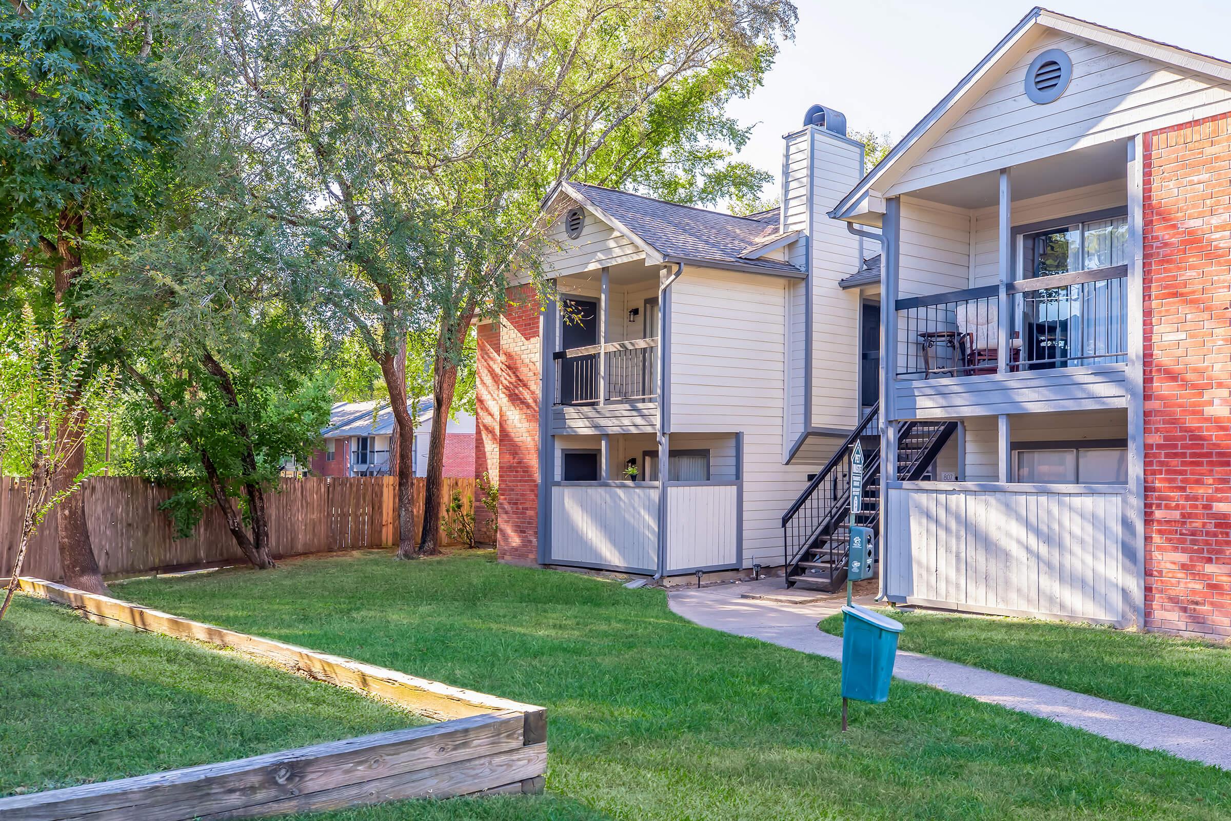 A two-story apartment building with a mix of light-colored and brick exterior. It features balconies, outdoor stairs, and a small green lawn with a garden bed. Trees provide shade, and a pathway leads through the grass. A trash bin is visible in the foreground.