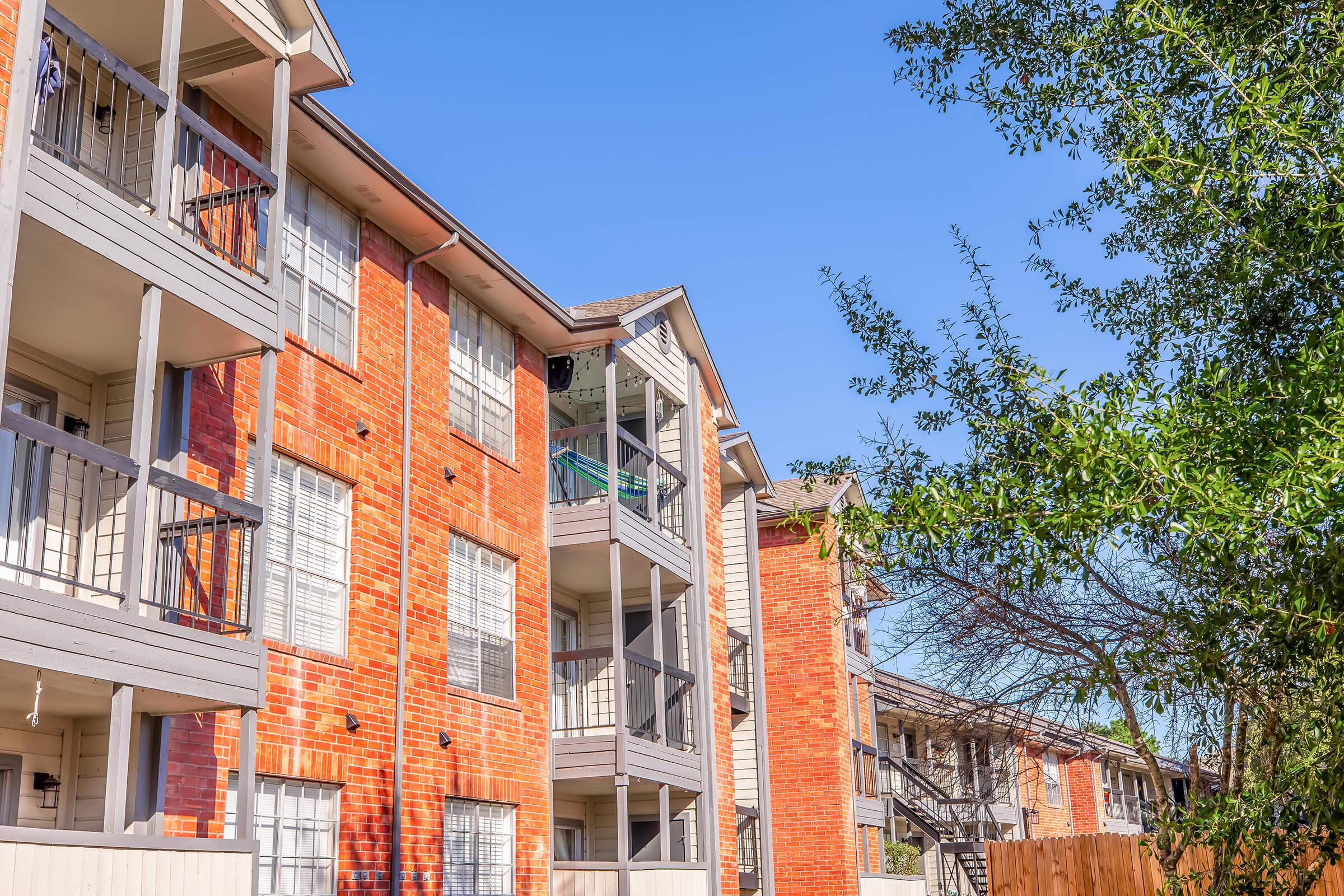 A sunny day view of a residential apartment complex with brick exteriors and balconies. The buildings feature multiple stories, and lush green foliage is visible in the foreground, creating a pleasant and inviting atmosphere. The blue sky enhances the vibrant colors of the architecture.