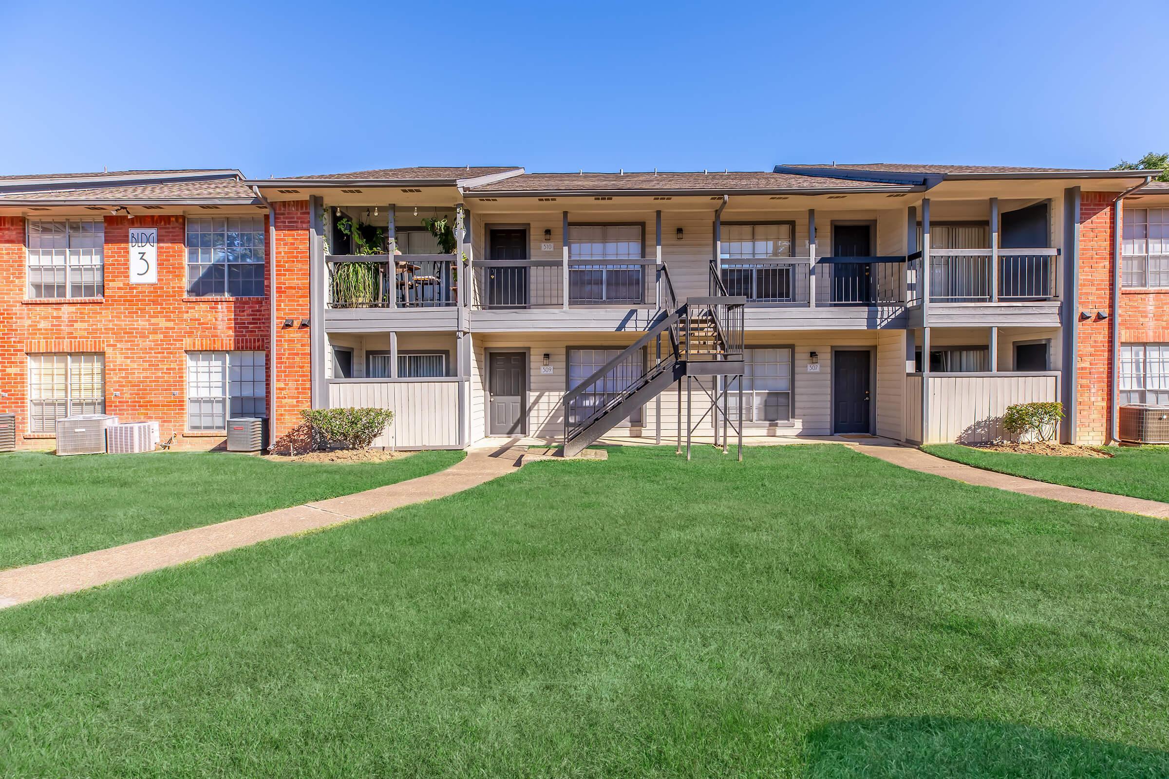 A landscaped courtyard featuring two multi-story brick apartment buildings. The buildings have balconies and staircases, with green grass and a pathway leading through the area. Bright blue sky above complements the sunny setting.