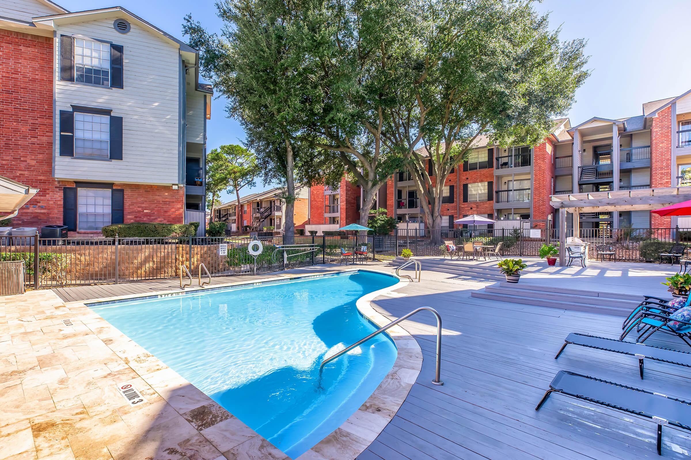 A sunny outdoor pool area surrounded by an apartment complex, featuring a curved pool, comfortable lounge chairs, and shaded umbrellas. Lush green trees provide shade, and the buildings are a mix of red brick and light-colored siding, creating a welcoming community atmosphere.