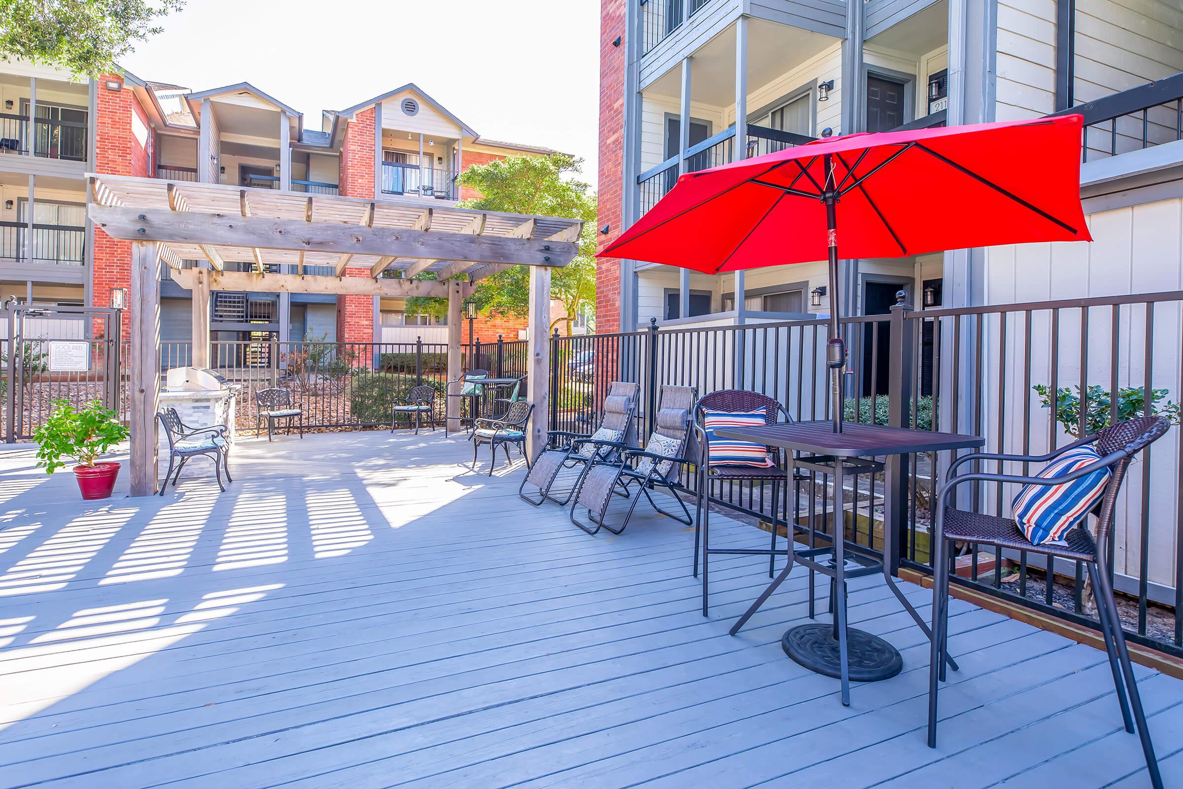 A spacious outdoor patio featuring a red umbrella over a small round table surrounded by chairs. There are additional seating options and a pergola in the background, with a view of an apartment complex. The deck is well-lit, creating a welcoming atmosphere.