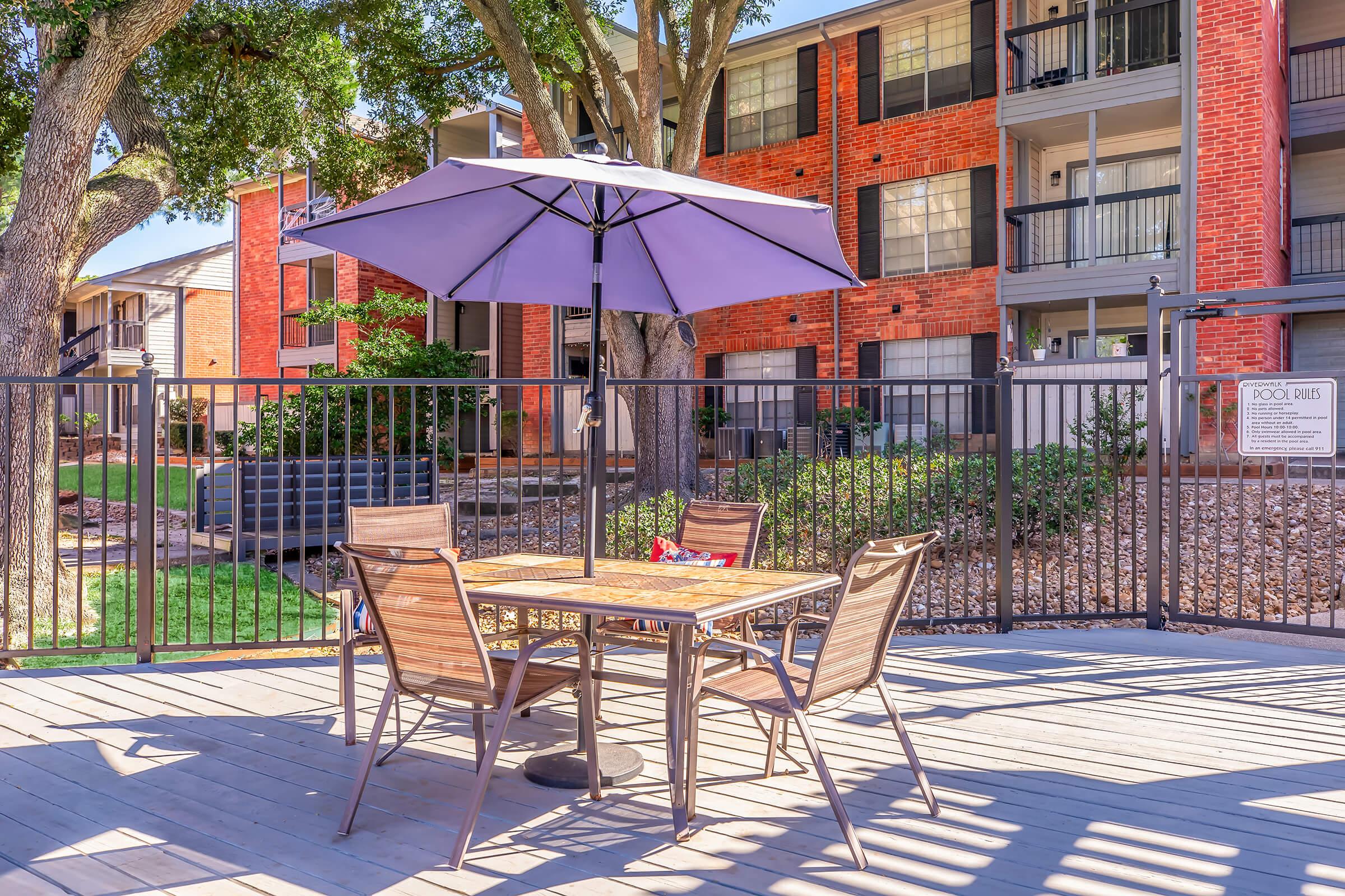 A patio area featuring a wooden table with four chairs under a purple patio umbrella. In the background, there are brick apartment buildings and a landscaped garden with trees. A fence encloses the space, creating a cozy outdoor setting for relaxation or dining.