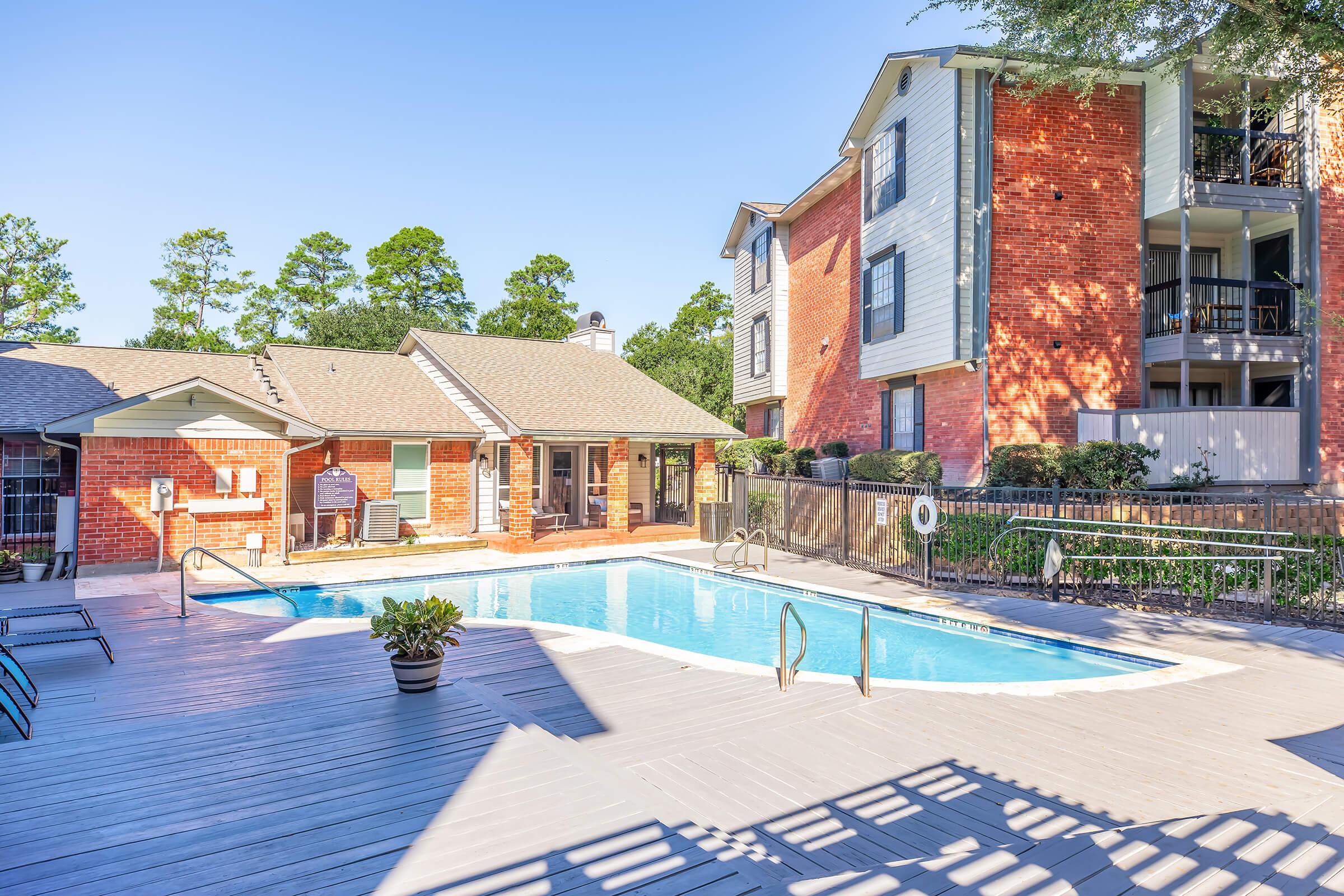A sunny outdoor swimming pool surrounded by wooden decking and lounge chairs. In the background, there are red brick apartment buildings with balconies and lush trees. The area appears well-maintained and inviting, perfect for relaxation and leisure activities.
