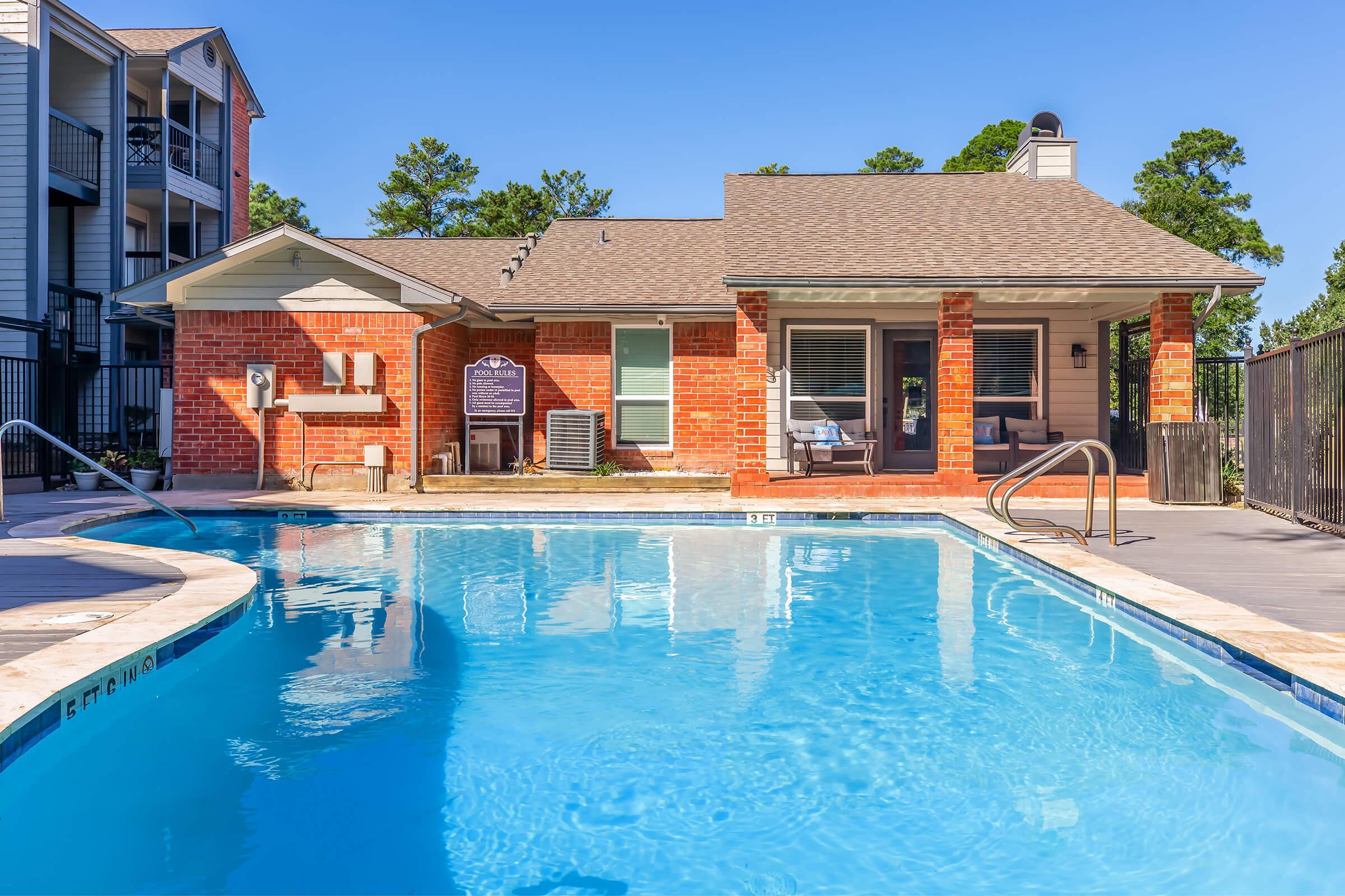 A bright, sunny view of a swimming pool surrounded by a patio area. The pool is crystal clear, with a ladder for easy access. In the background, there is a brick building with large windows and a covered porch, surrounded by trees. The sky is clear and blue, adding to the inviting atmosphere.