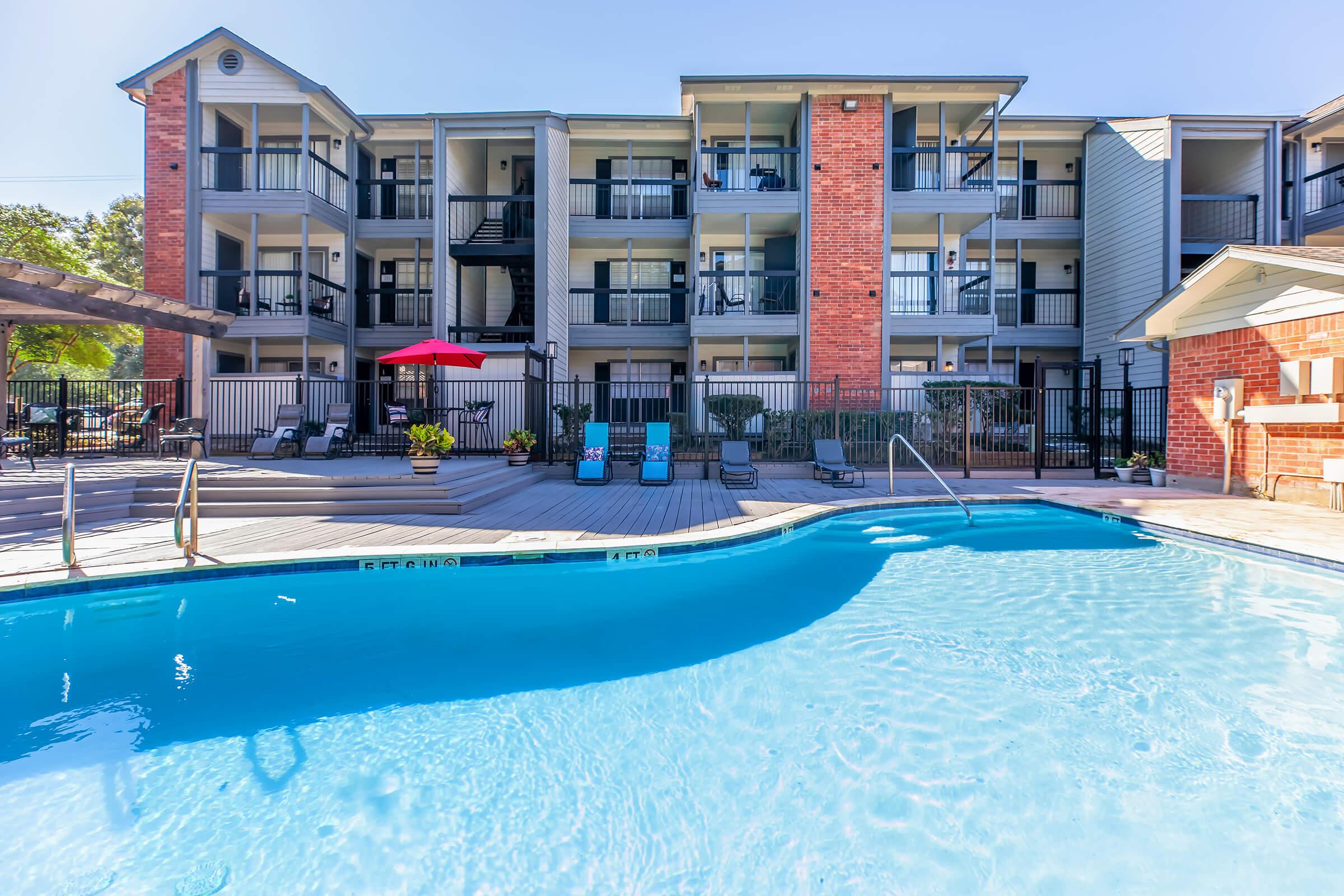 A clear blue swimming pool in the foreground, surrounded by wooden decking and lounge chairs. In the background, a three-story apartment building features balconies and a red umbrella near the pool area. Lush greenery is visible around the property, creating a welcoming outdoor space.