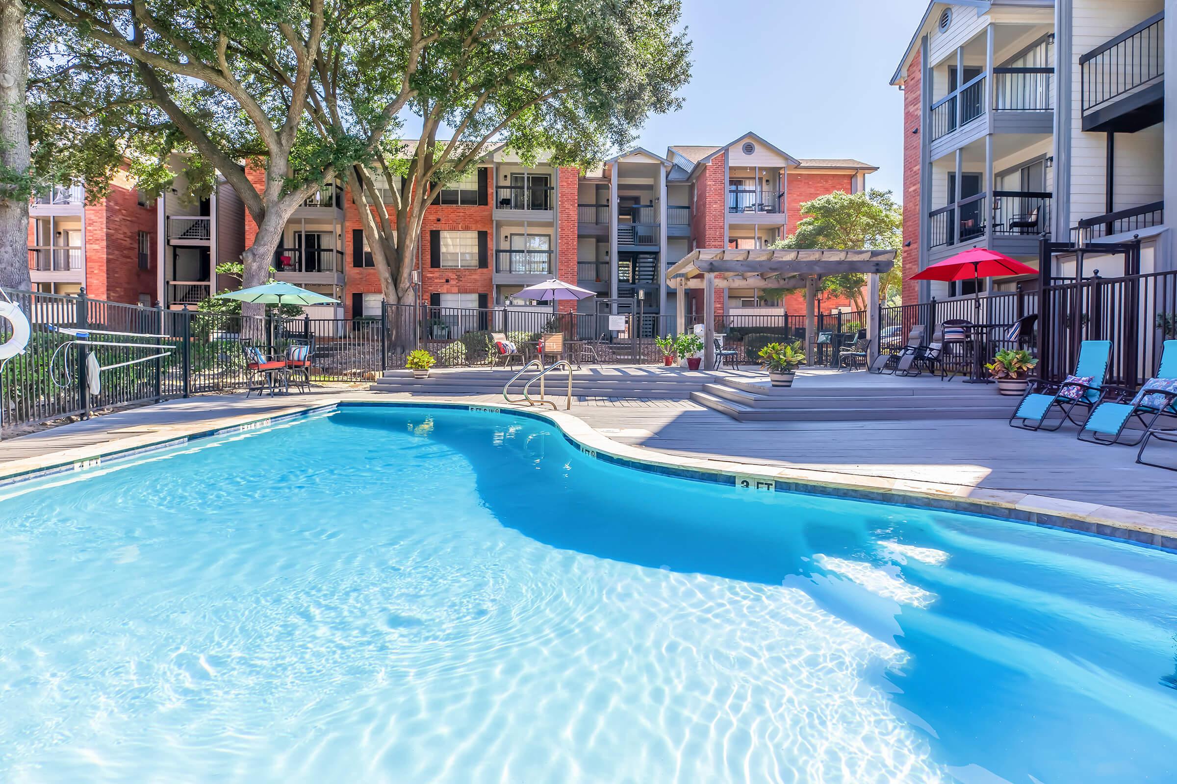 A bright, inviting swimming pool surrounded by a wooden deck, lounge chairs, and colorful umbrellas. In the background, there are apartment buildings with balconies and lush trees providing shade, creating a relaxing outdoor atmosphere.