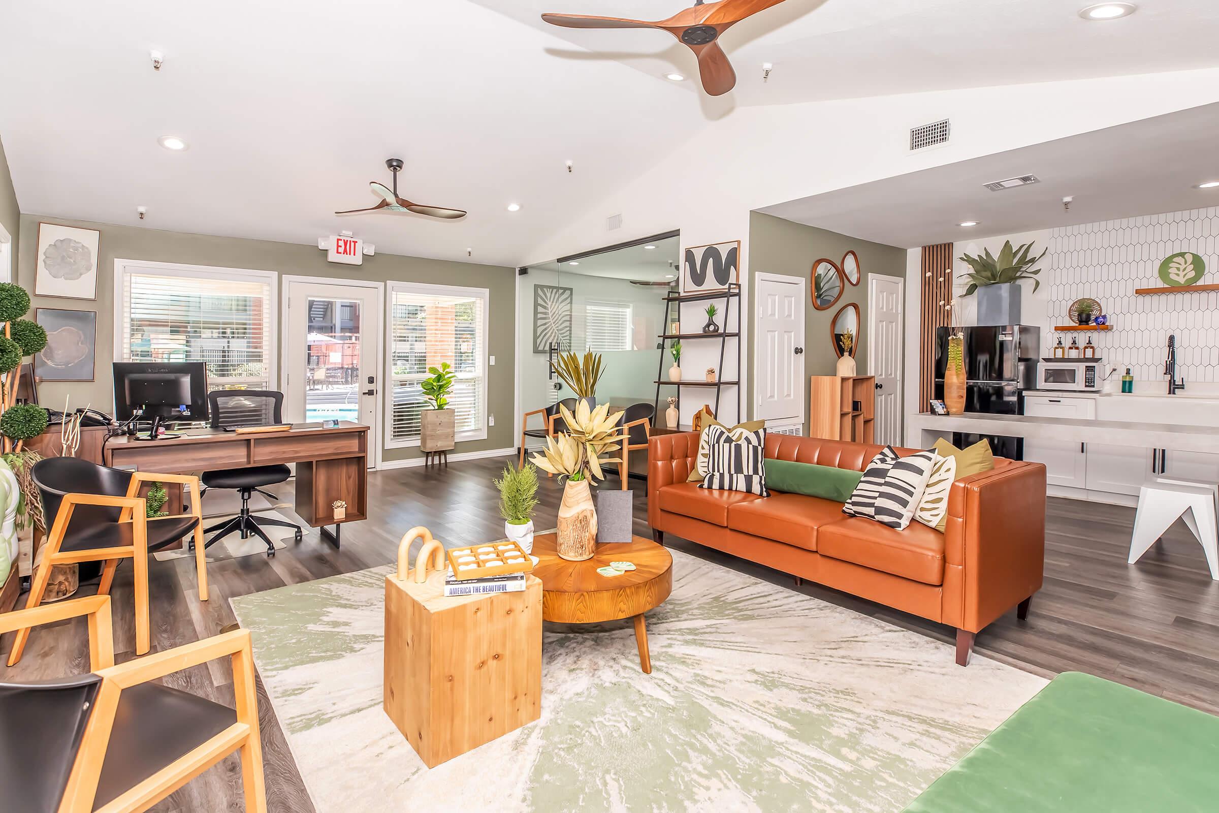 Modern lounge area featuring a mix of comfortable seating, including a brown leather sofa, wooden chairs, and a coffee table. The space is decorated with plants and artwork, along with a workspace that has a computer. Large windows let in natural light, creating a welcoming atmosphere.