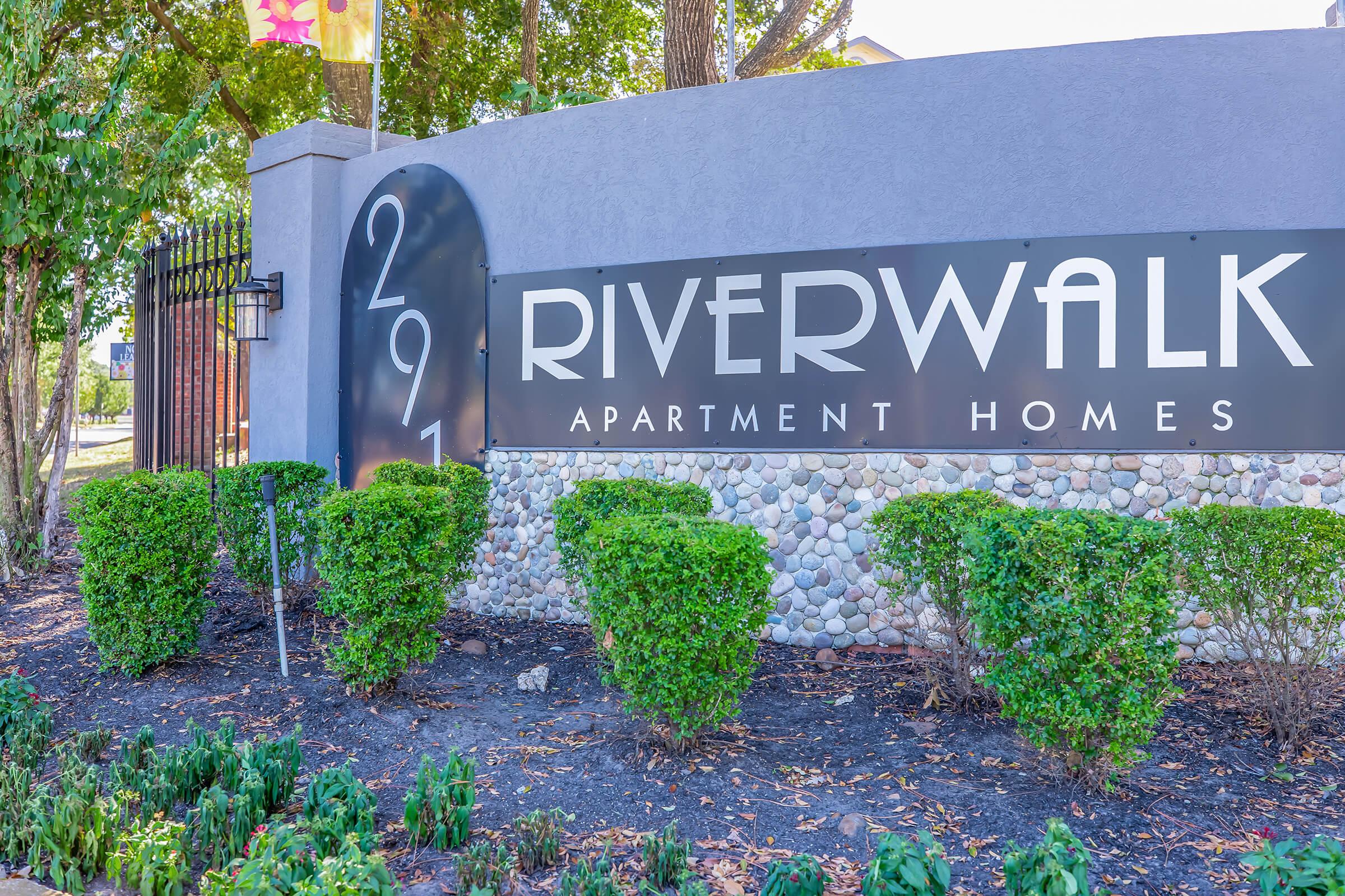 Sign for Riverwalk Apartment Homes featuring the number 291, surrounded by neatly trimmed bushes and landscaping. The sign is prominent against a grey background, with a decorative fence and trees in the vicinity.