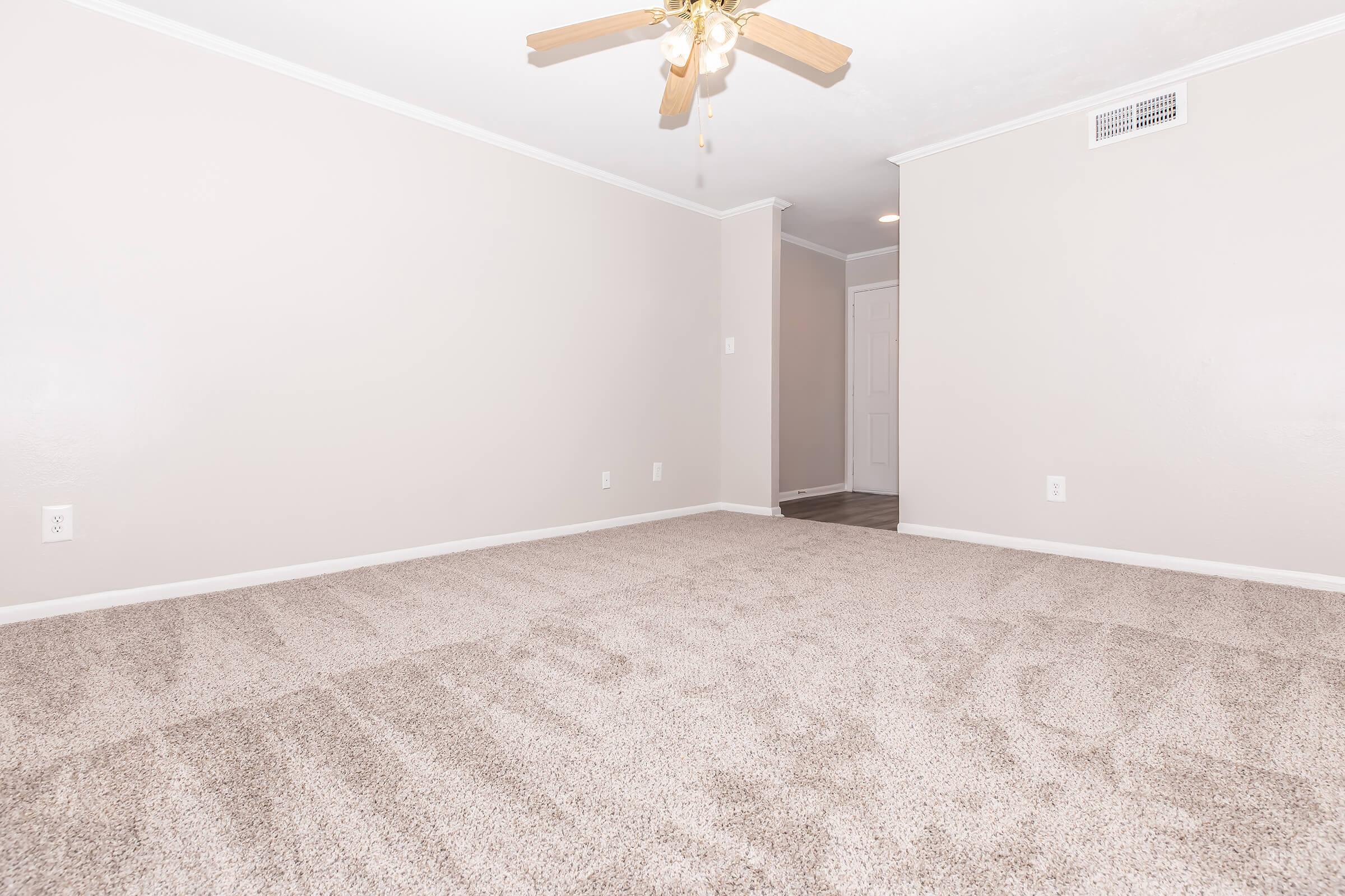 A spacious room with light-colored walls and a beige carpet. The ceiling features a ceiling fan with wooden blades. A doorway is visible in the background, leading to another room. The overall ambiance is bright and neutral, with no furniture present.