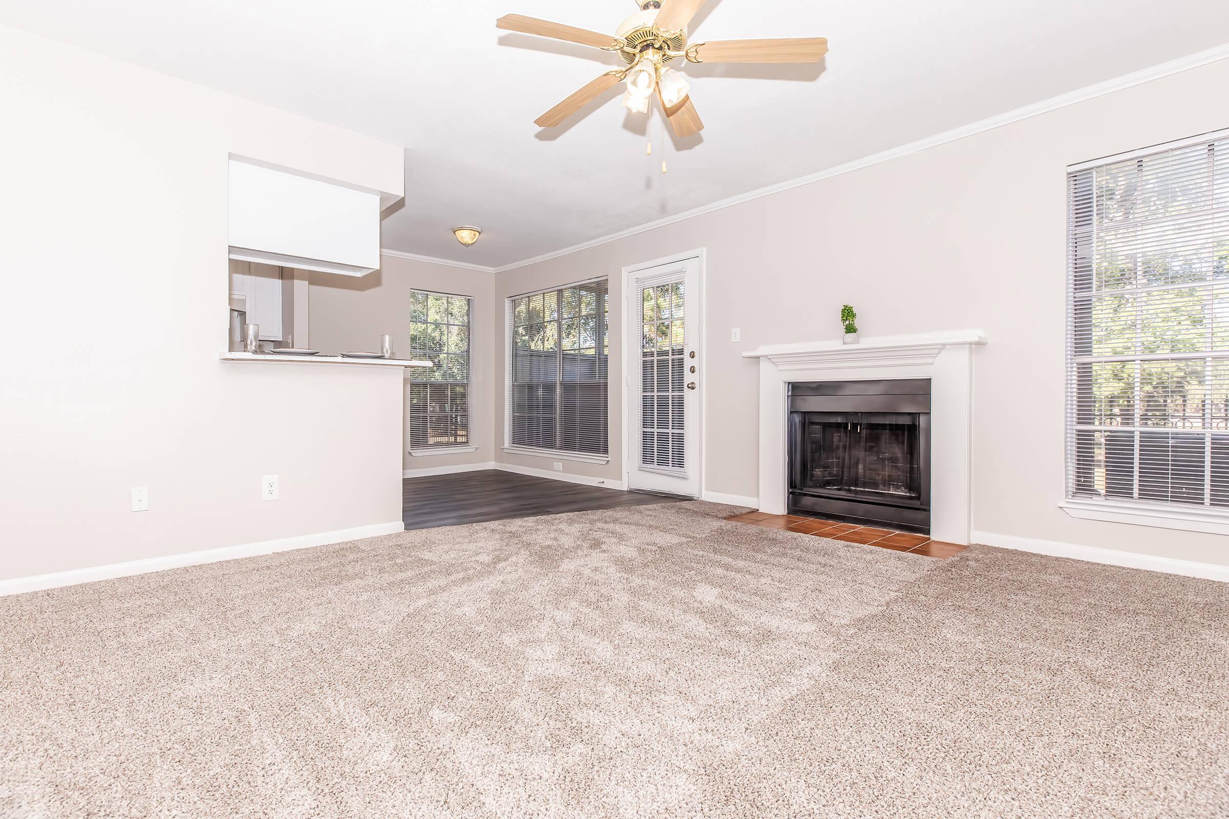 A spacious living room with light-colored walls and a ceiling fan. The floor is carpeted, and there's a cozy fireplace with a white mantel. Large windows let in natural light, and there's a sliding door leading outside. The area is open to a kitchen space in the background.