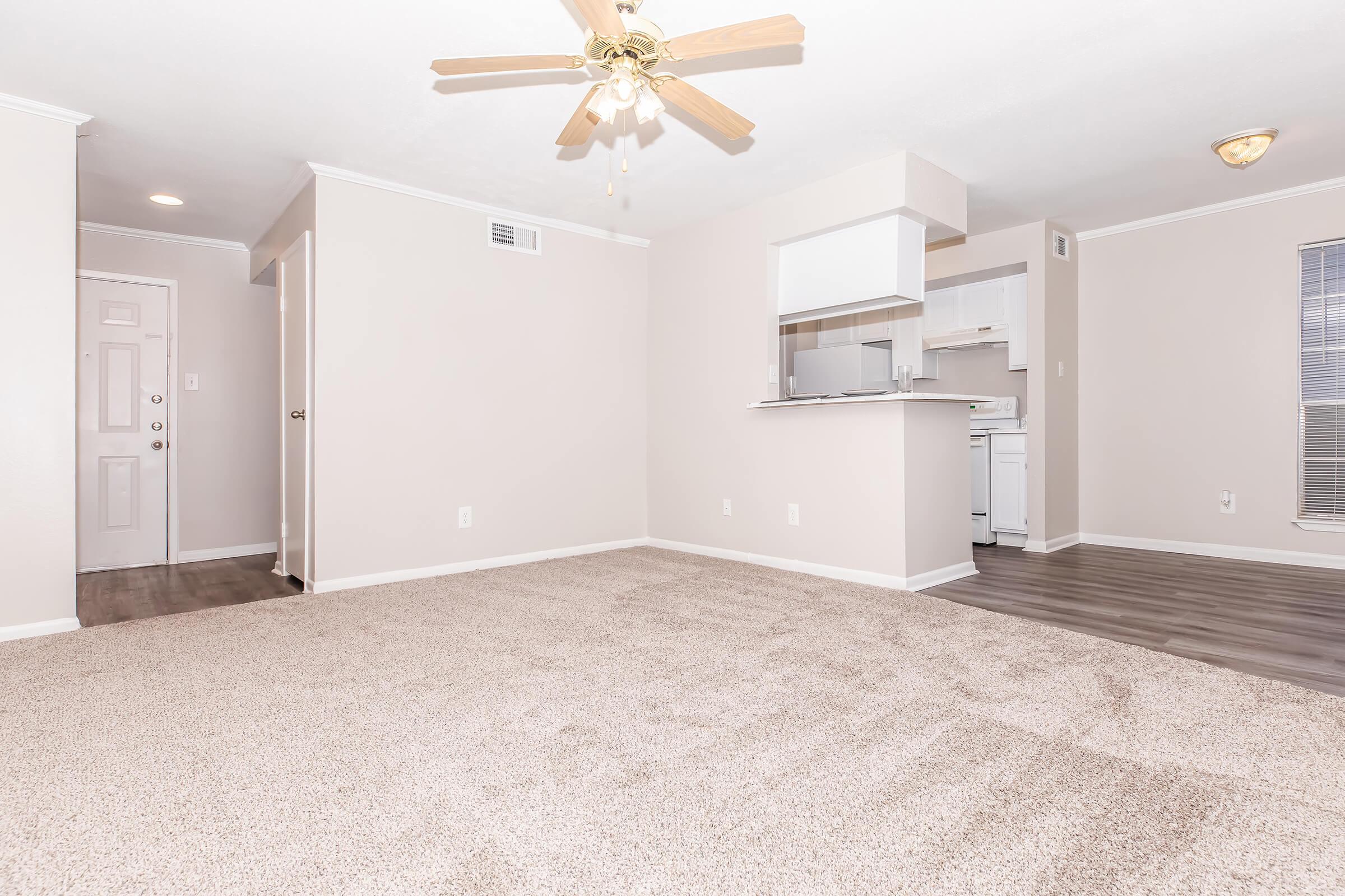 A spacious and clean living area with light beige walls and plush carpet. A ceiling fan hangs above, and there is a doorway leading to another room. In the background, a small kitchen area is visible with white cabinetry, and a door leads to the outside. Natural light enters through a window.