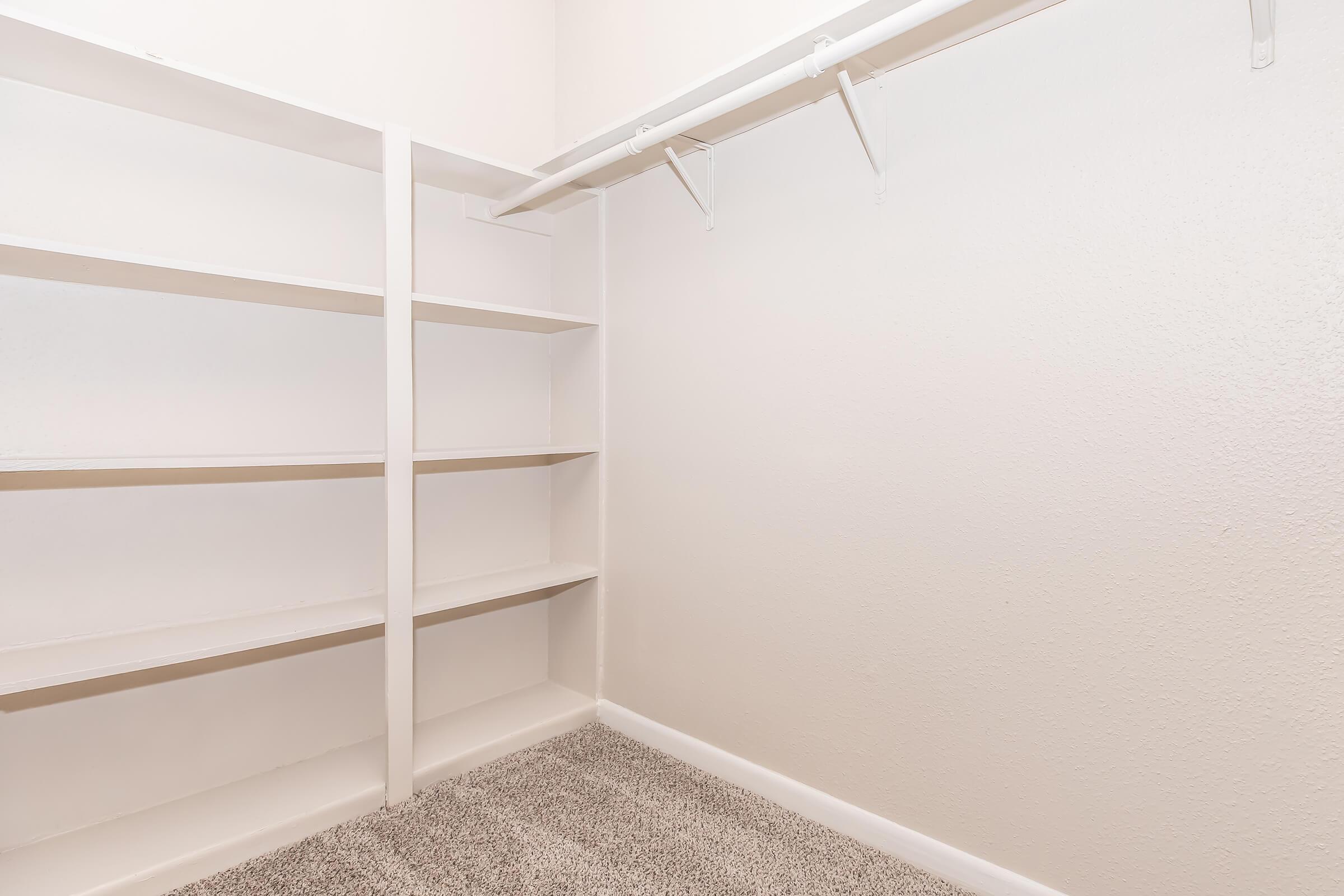 A small, empty closet featuring white shelving units and a hanging rod. The walls are light-colored and the floor is carpeted. The space is well-lit and appears tidy, with no items or decorations present.