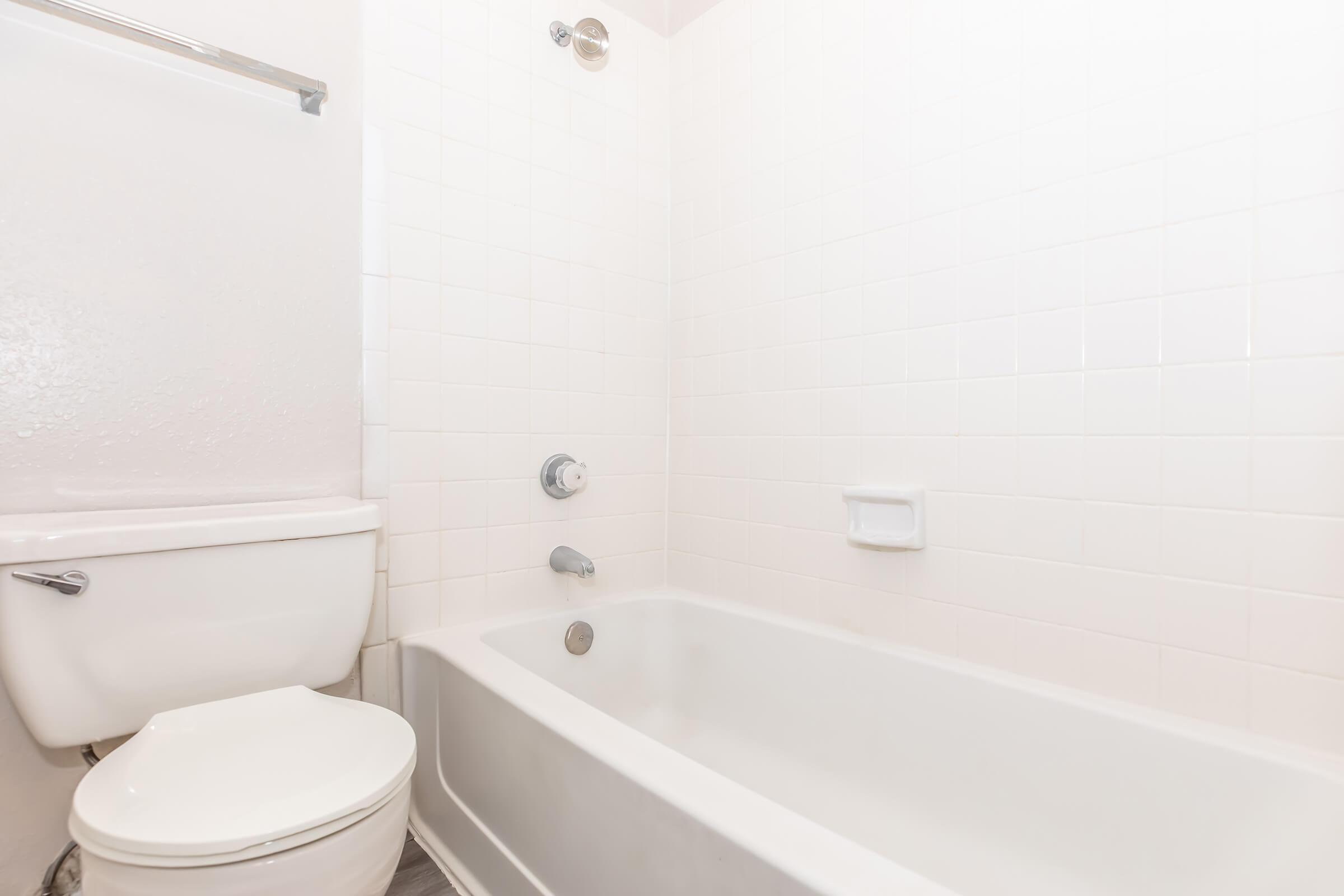 A clean, light-colored bathroom featuring a white bathtub, a wall-mounted faucet, and a toilet. The walls are tiled in white, creating a bright and spacious feel. The floor is visible, adding to the simplicity of the design.