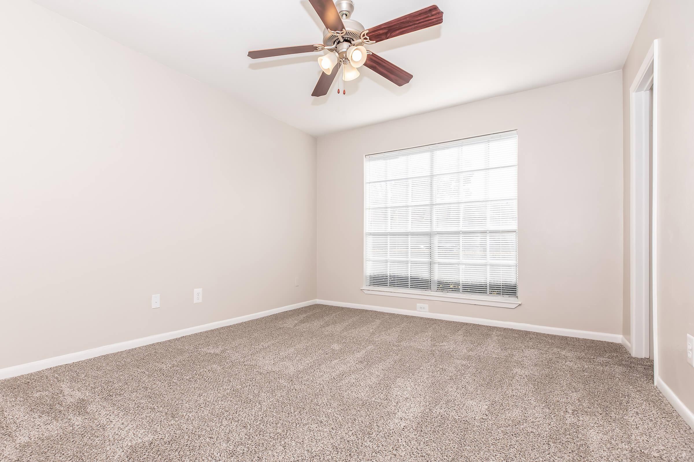 A spacious, empty room with beige walls and a light-colored carpet. A ceiling fan with five wooden blades hangs above, and a large window with white blinds allows natural light to enter. The room has a clean and minimalistic appearance, suitable for various uses or decorating.