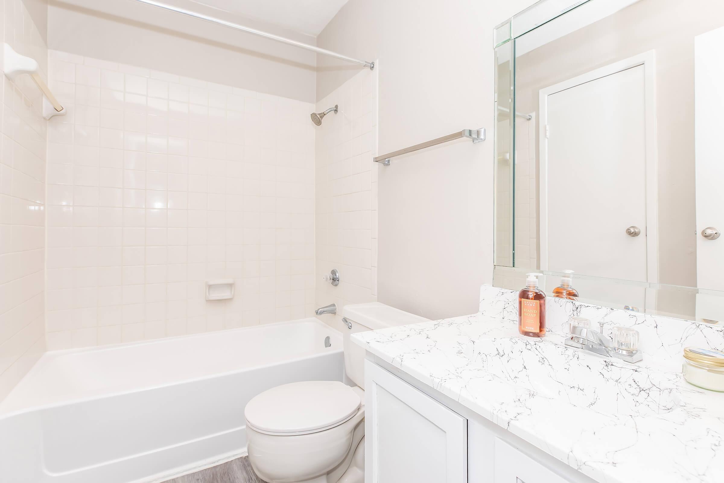 Clean, modern bathroom featuring a white bathtub and shower combination, a white toilet, and a stylish vanity with a marble countertop. The walls are light-colored, and there is a large mirror above the sink. Shelving is visible, along with two bottles on the countertop. Natural light illuminates the space.