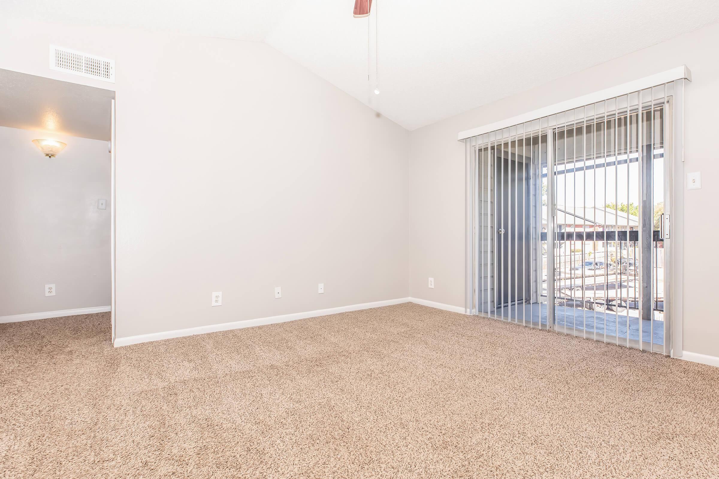 Spacious, empty living room with light beige carpet and neutral walls. A sliding glass door leads to a balcony, providing natural light. The room features a ceiling fan and an entrance to another space on the left.