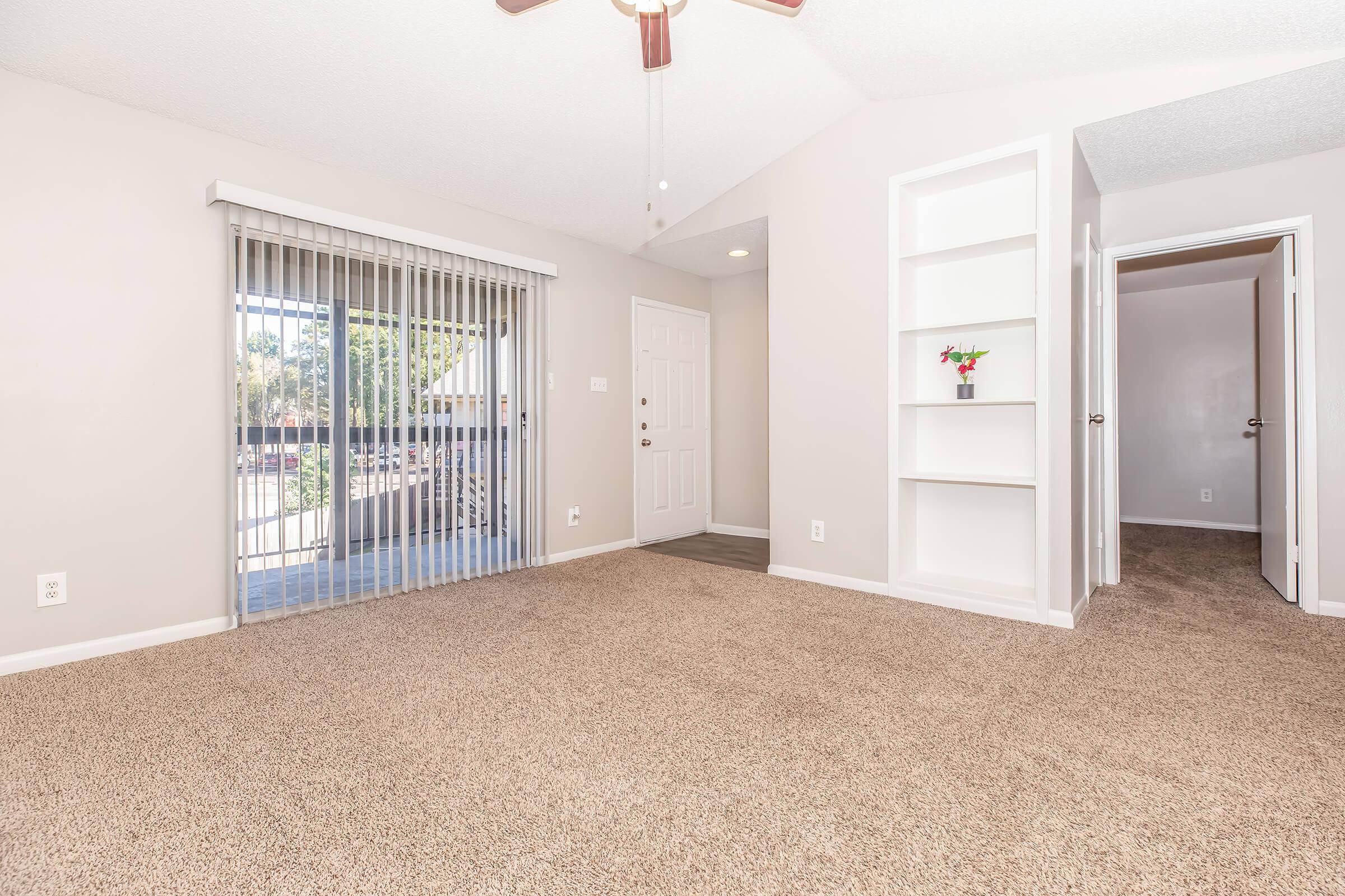 An empty living room with tan carpet, light-colored walls, and a ceiling fan. There's a sliding glass door leading to a balcony, along with a small decorative shelf and an open door on the right leading to another room. Natural light illuminates the space, creating a bright and airy atmosphere.