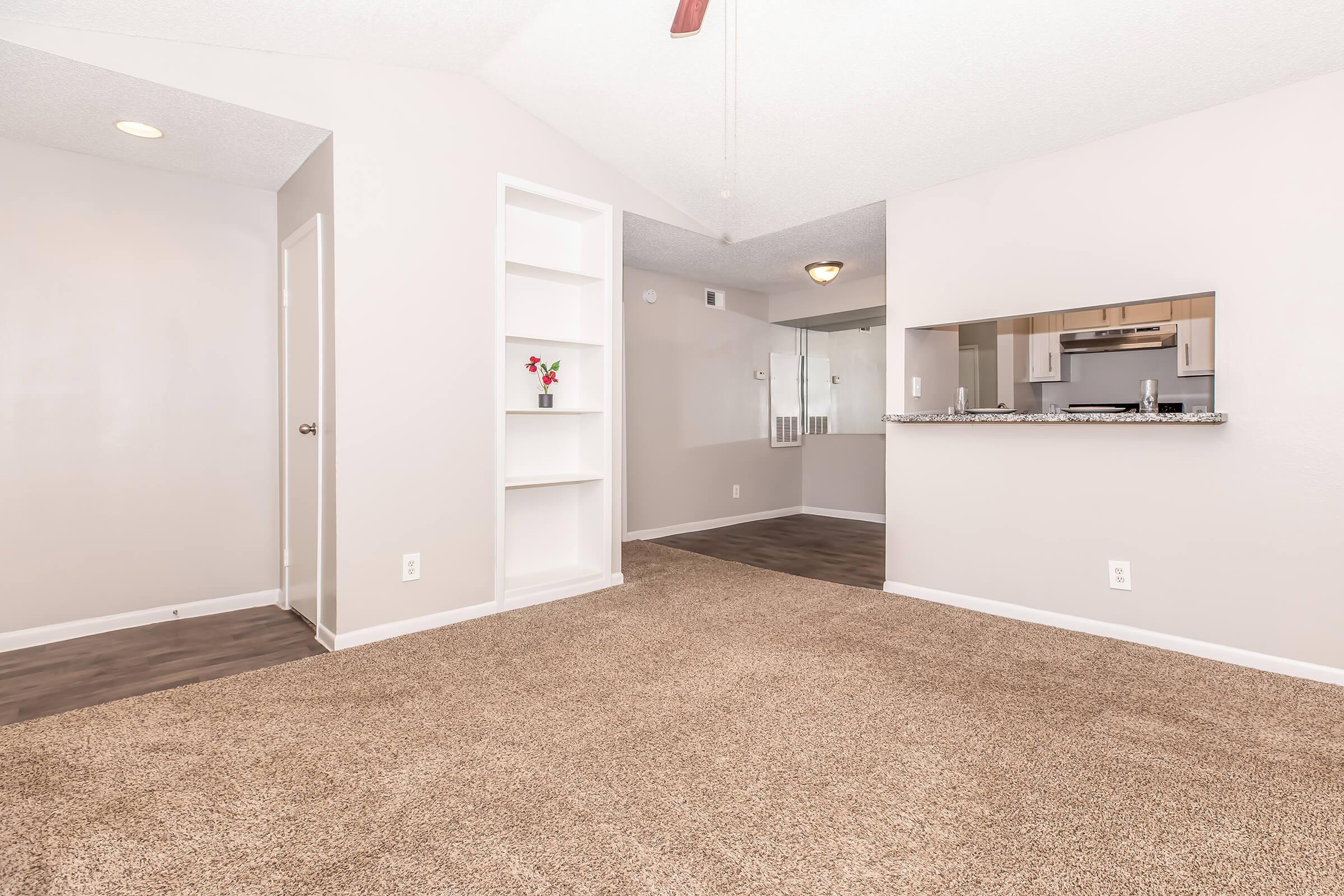 A spacious room featuring light-colored walls and carpet, with a ceiling fan and a small bookshelf in the corner. An open area connects to a kitchen with a bar counter. Natural light flows in, creating a welcoming atmosphere.
