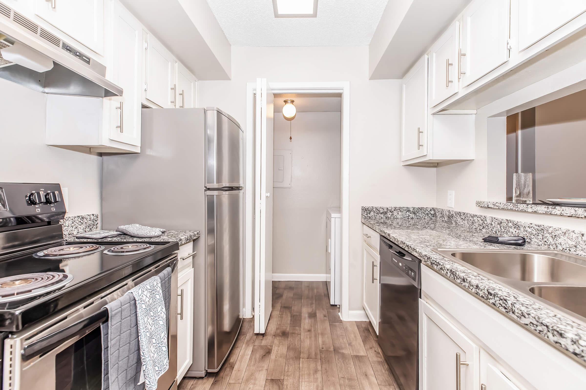 Modern kitchen featuring white cabinets, stainless steel appliances, and granite countertops. Includes an oven, stove, refrigerator, and dishwasher. The space is well-lit with natural light and has a central ceiling fan, with a doorway leading to a hallway and additional rooms.