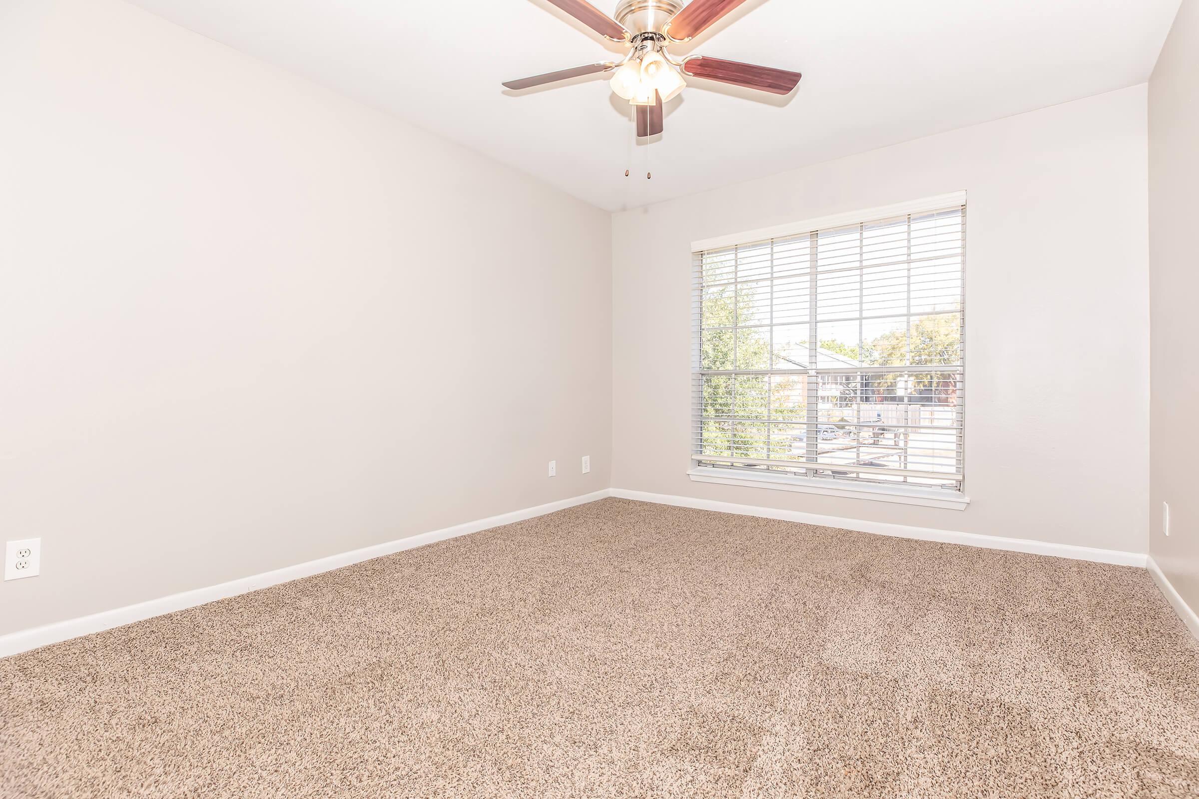 A spacious, empty room featuring beige walls and plush carpet. A ceiling fan is mounted in the center, and a large window with blinds allows natural light to enter. The view outside shows trees and a clear sky, creating a bright and inviting atmosphere.