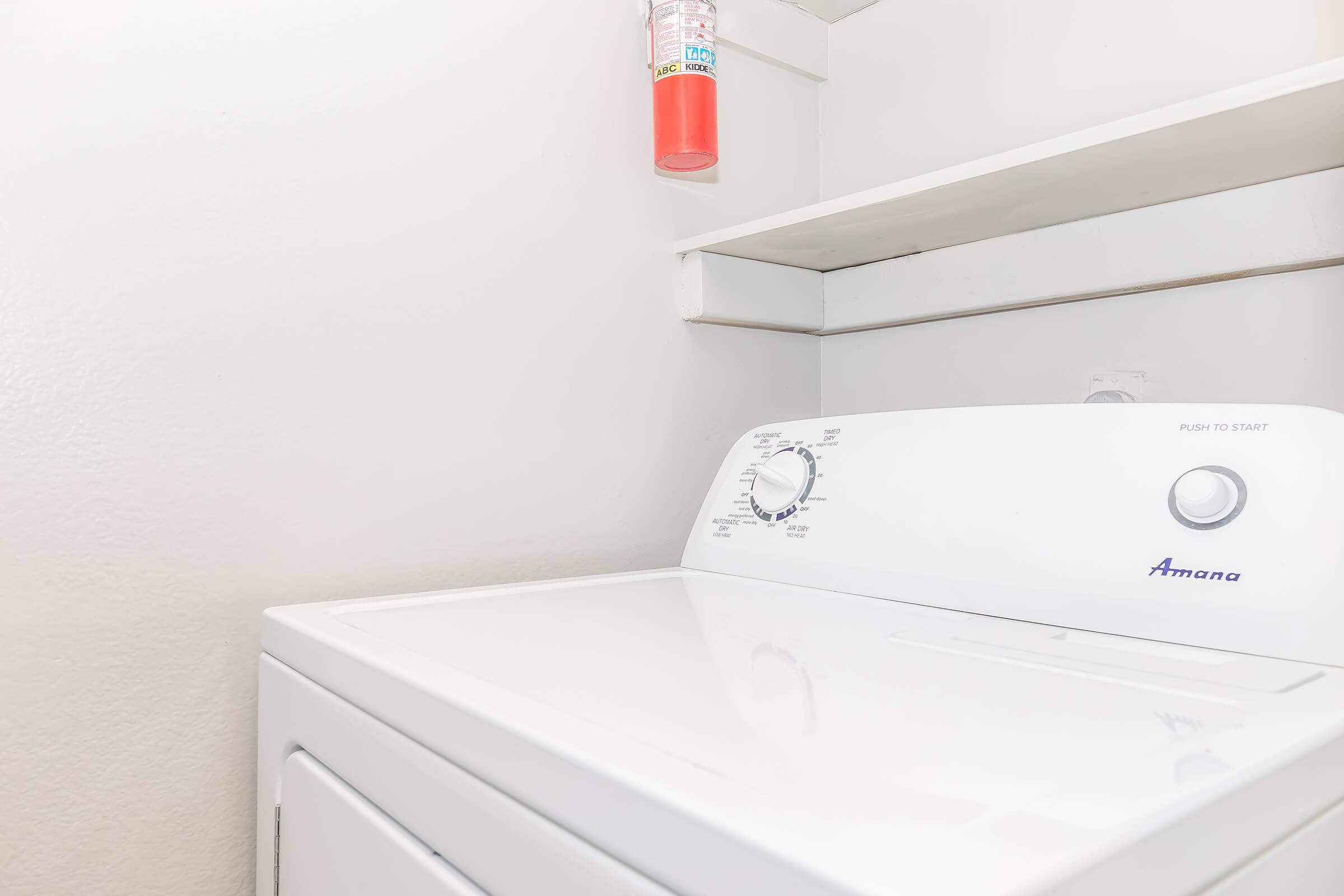 A white Amana washing machine with control knobs is positioned against a light-colored wall. Above the machine, there is a shelf holding a fire extinguisher. The setting appears clean and well-organized, typically found in a laundry room.
