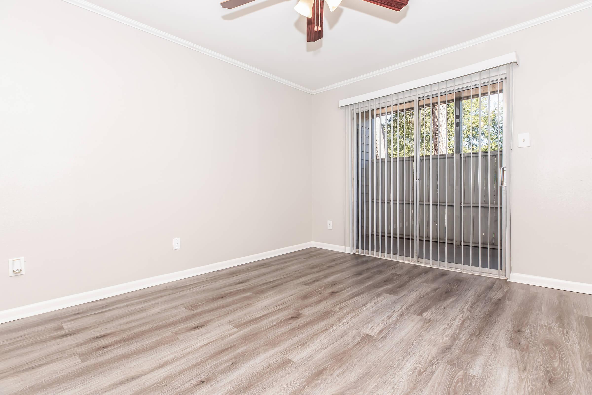 Empty room with light gray walls and a wooden floor. A ceiling fan is visible, and there are vertical blinds covering a large window that lets in natural light. The space appears spacious and well-lit, with no furniture or decorations present.
