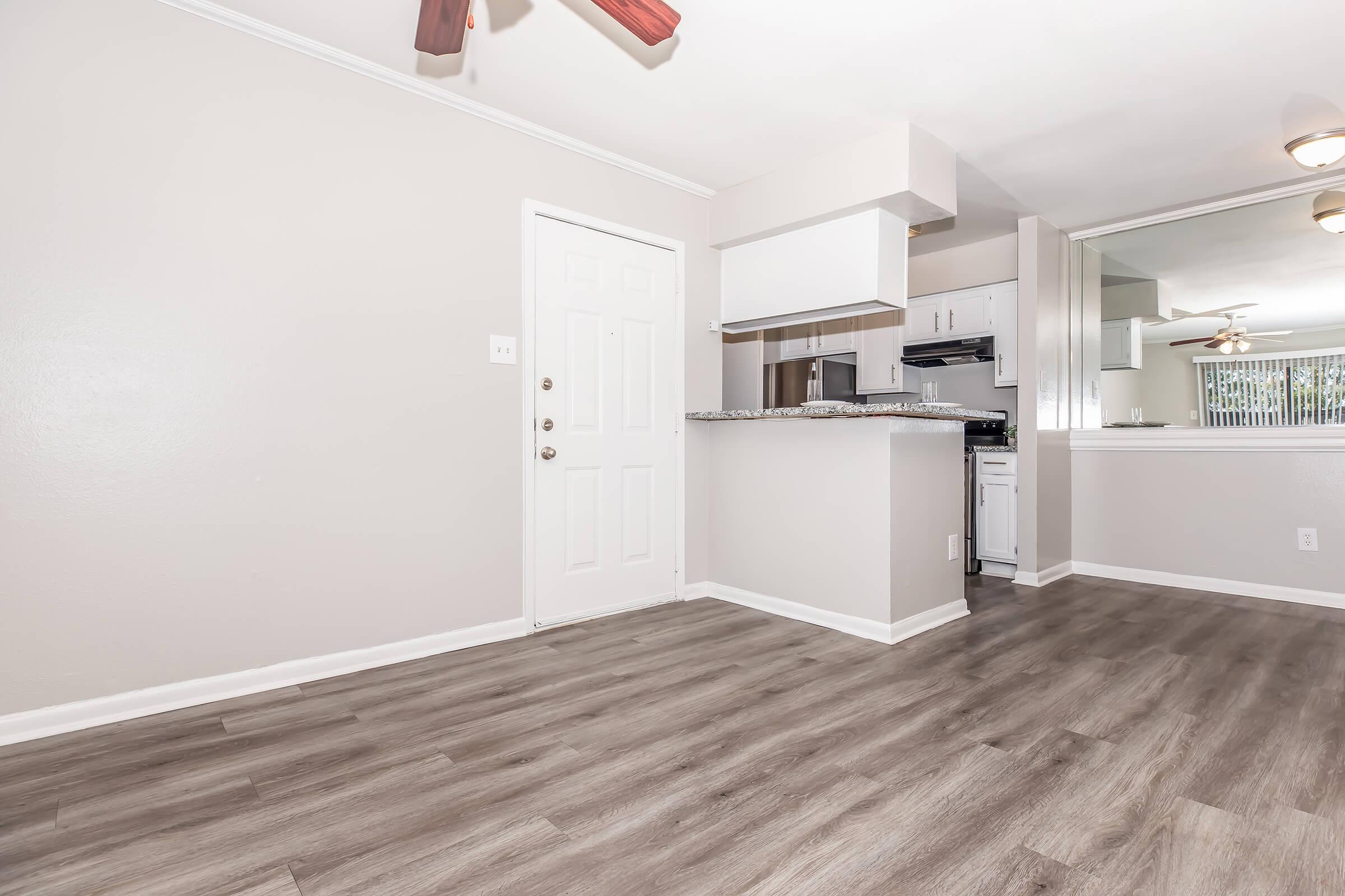 Interior view of a modern living space featuring light gray walls, a white front door, and new wood flooring. The kitchen area is visible with a small bar, white cabinets, and stainless-steel appliances. Large windows provide natural light, creating an airy atmosphere.