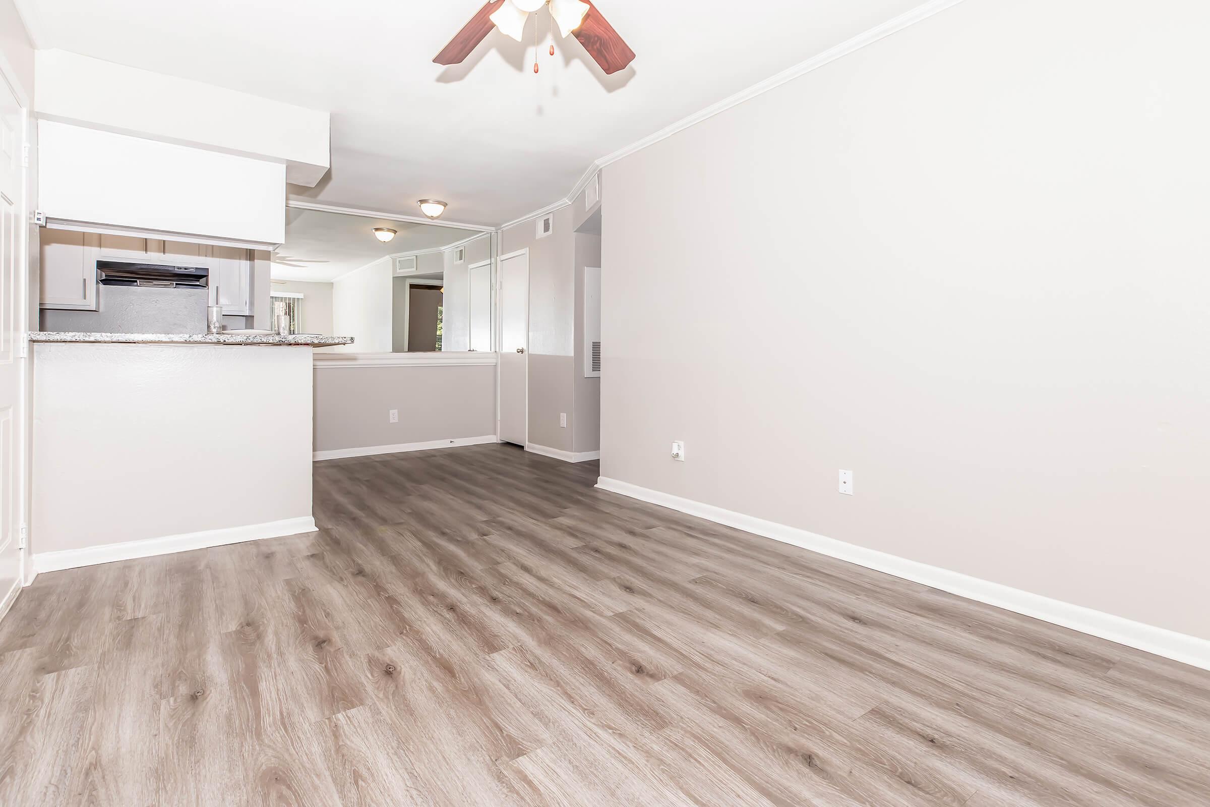 Spacious living area with light-colored walls and modern laminate flooring. A ceiling fan hangs above, and an open kitchen area is visible in the background. Natural light filters through a nearby window, creating a bright and inviting atmosphere.