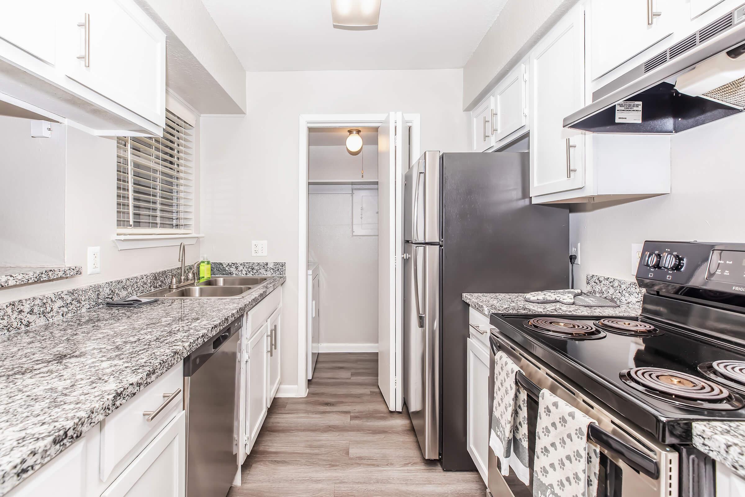 Modern kitchen featuring granite countertops, stainless steel appliances, and white cabinetry. The space includes a double sink, an electric stove with an oven, and a refrigerator. Light-colored walls and wooden flooring create a bright and inviting atmosphere. A doorway leads to an adjacent room.