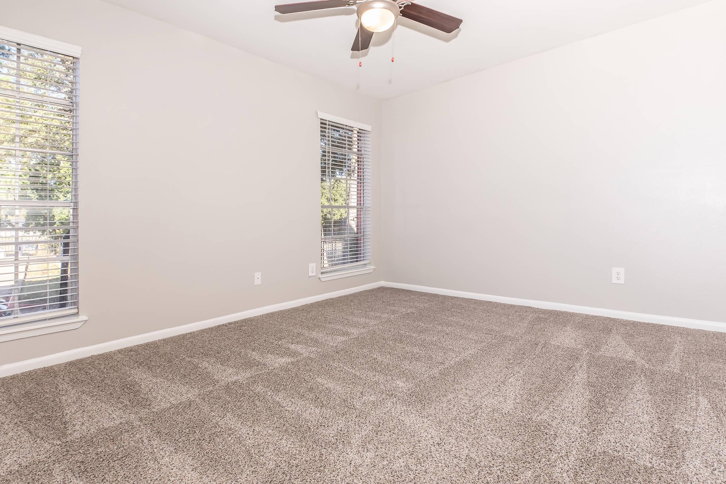 A spacious, empty room with light gray walls and a light carpeted floor. It features two windows with blinds letting in natural light and a ceiling fan overhead. The overall atmosphere is bright and inviting, perfect for personal customization.