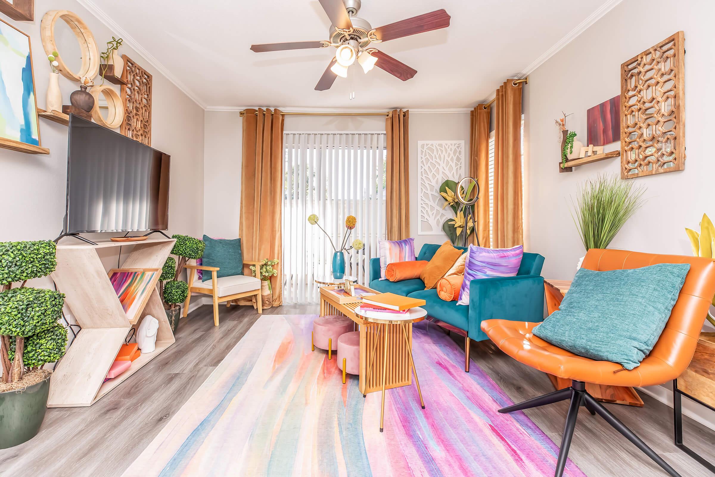 Bright and modern living room featuring a teal sofa with colorful throw pillows, an orange accent chair, and a wooden coffee table. A large TV is mounted on the wall, complemented by decorative plants and art on the walls. Natural light streams through sheer curtains, creating a warm and inviting atmosphere.